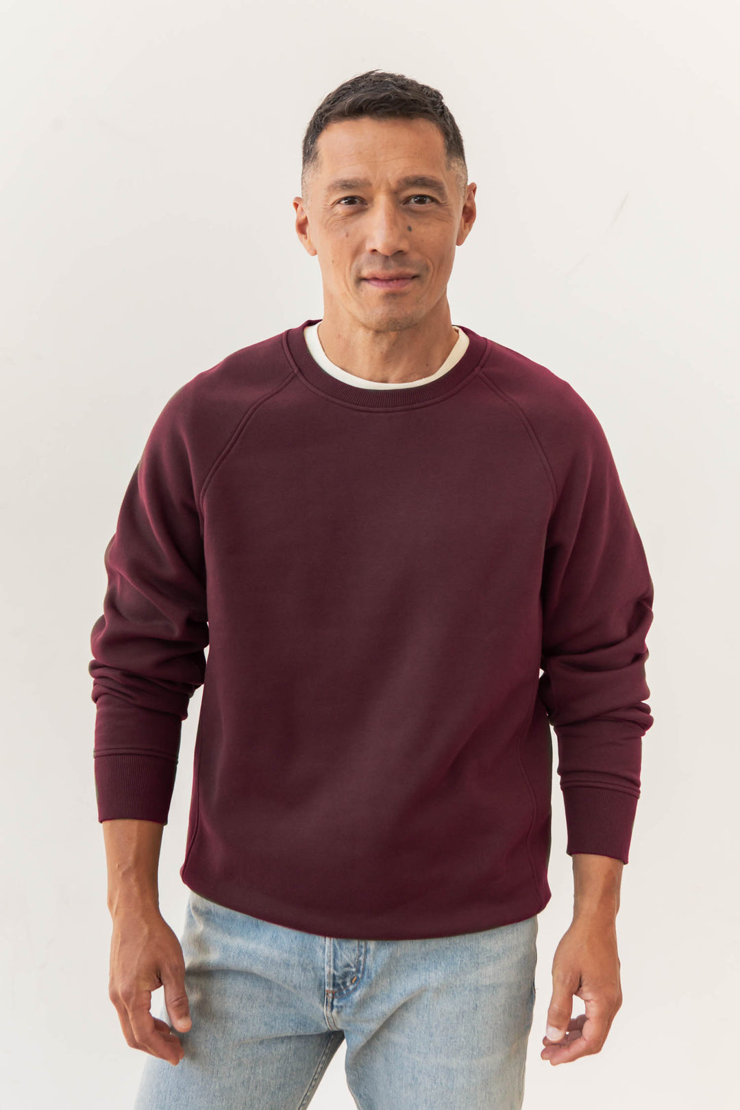 A man with short dark hair wears the Bridge & Burn Lincoln Crew Sweatshirt in Burgundy and light blue jeans, standing against a plain white background, facing the camera with a slight smile.