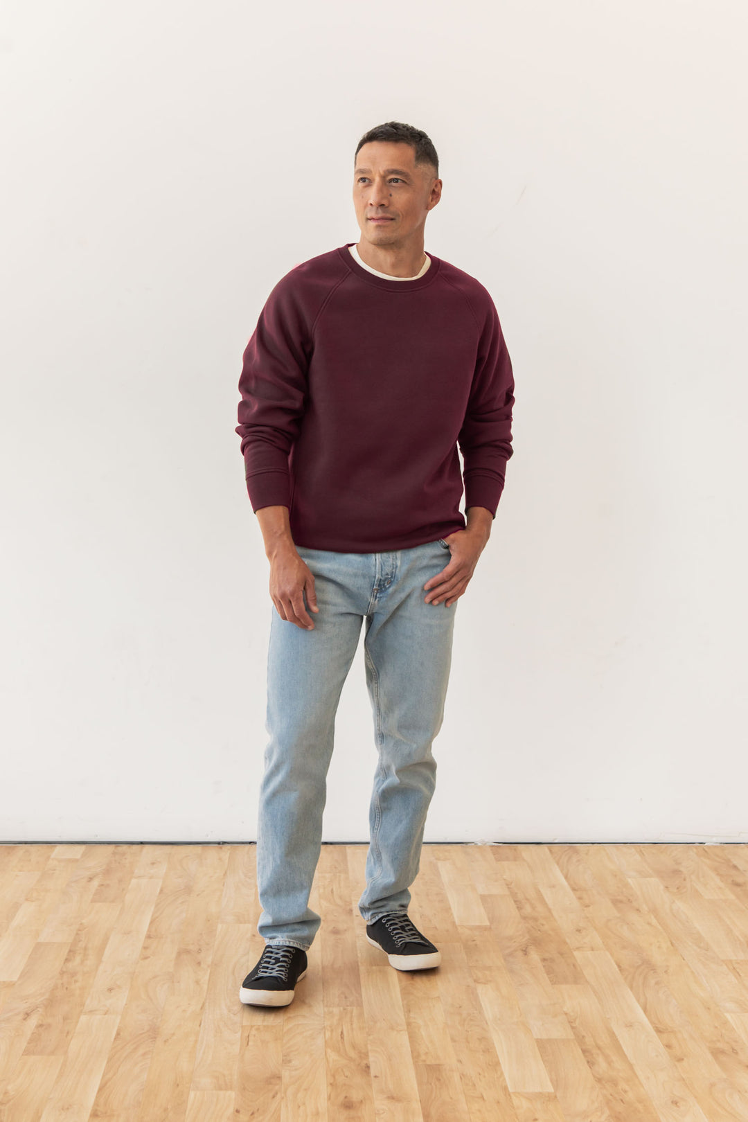 A man stands on a wooden floor against a white wall, wearing the Lincoln Crew Sweatshirt in Burgundy by Bridge & Burn, light blue jeans, and black sneakers. He looks slightly to the side with one hand in his pocket.