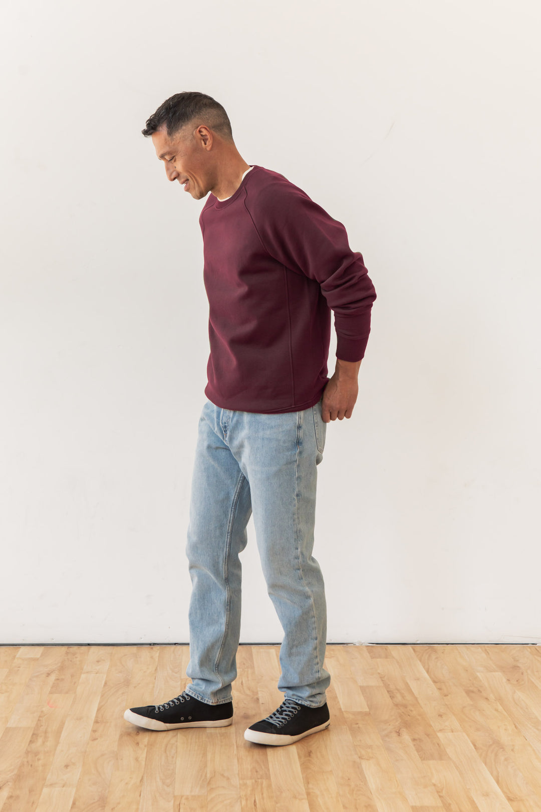 A man wearing a Bridge & Burn Lincoln Crew Sweatshirt in burgundy, light blue jeans, and black sneakers stands on a wooden floor against a plain white wall, looking down and smiling.