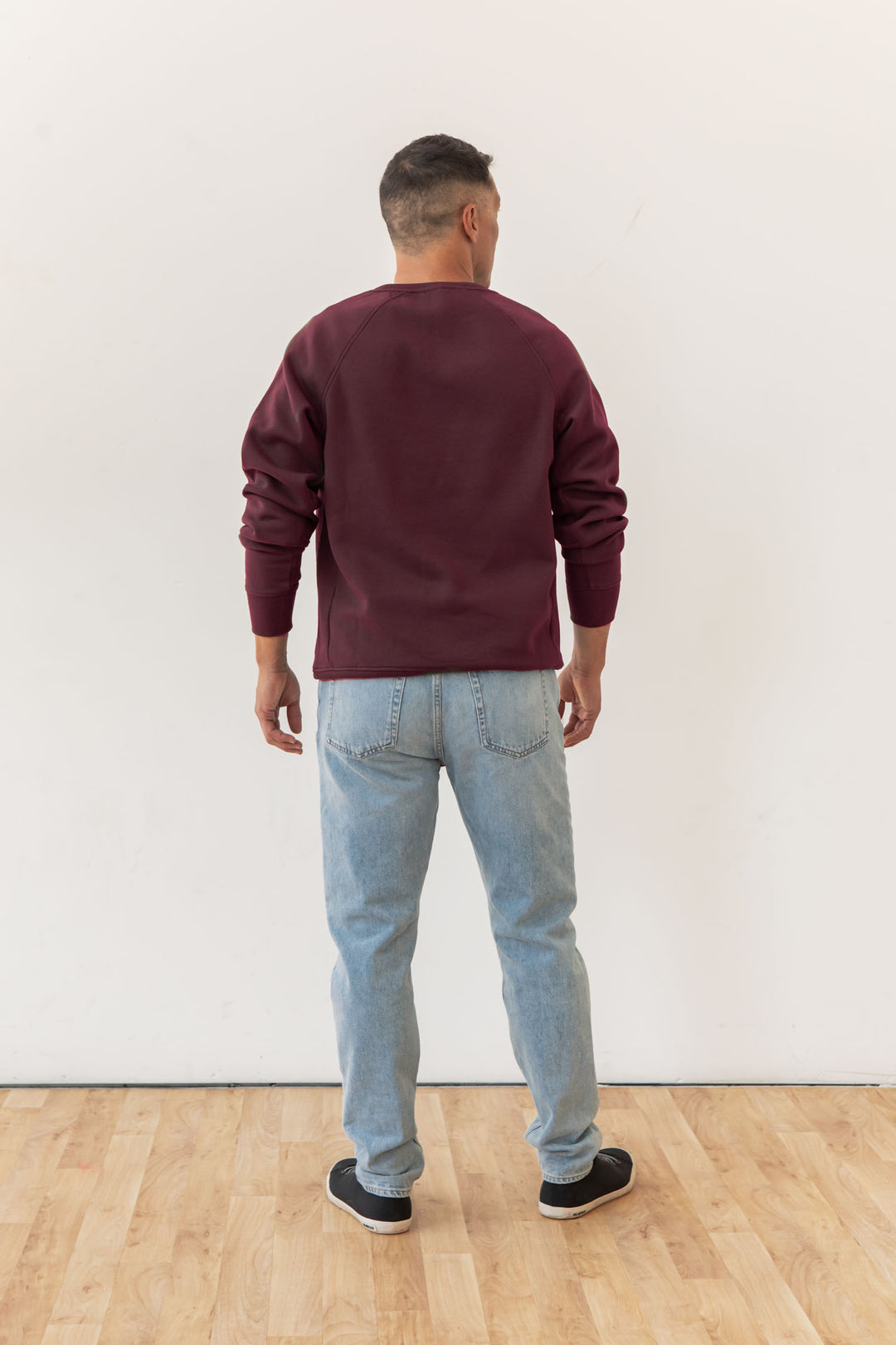 A man stands with his back to the camera, wearing the Bridge & Burn Lincoln Crew Sweatshirt in Burgundy, light blue jeans, and black sneakers on a light wooden floor against a plain white wall.
