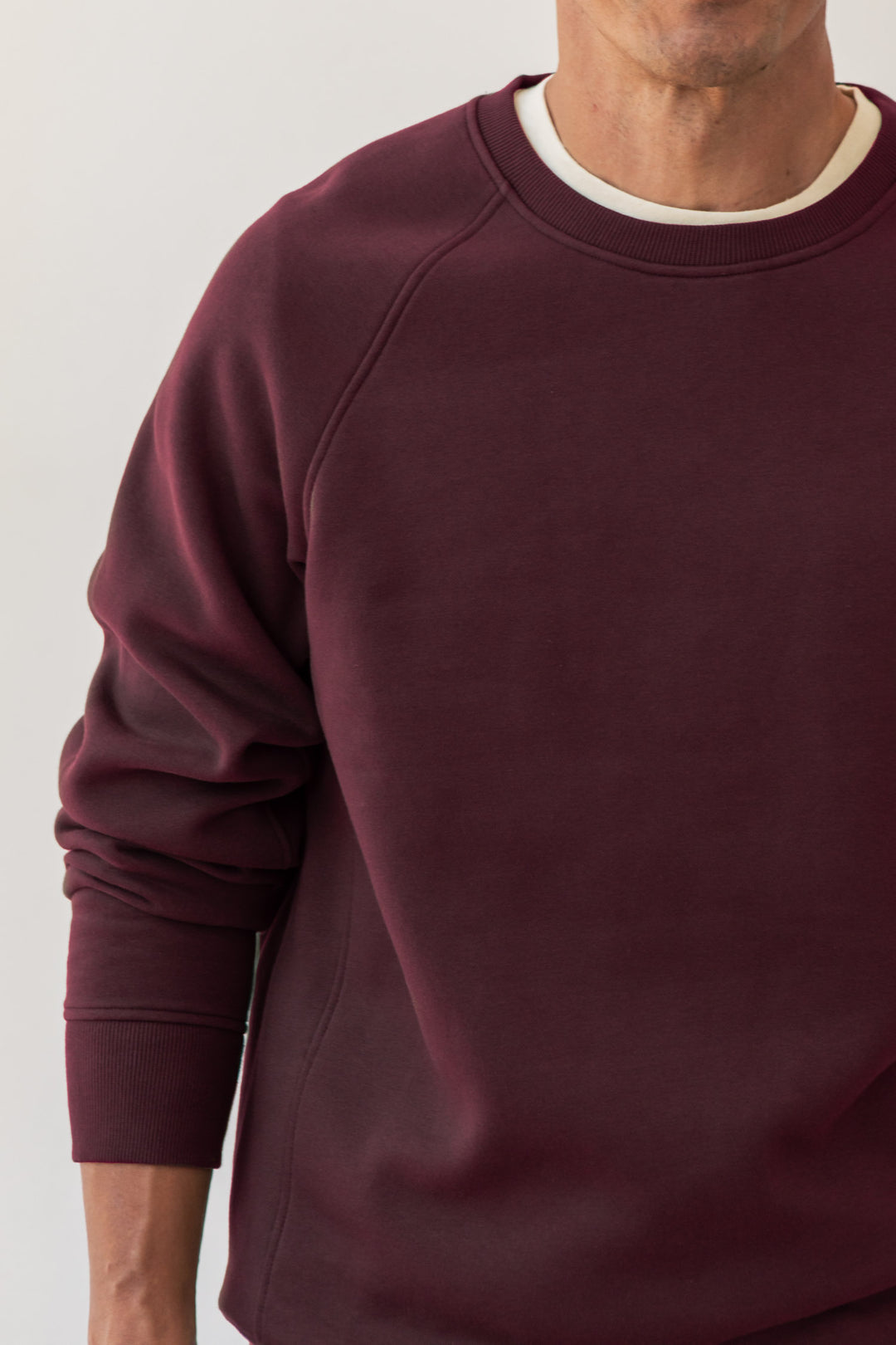 A person wearing the Bridge & Burn Lincoln Crew Sweatshirt in Burgundy over a white undershirt, shown from shoulders to waist, stands against a light background with their face partially out of frame.