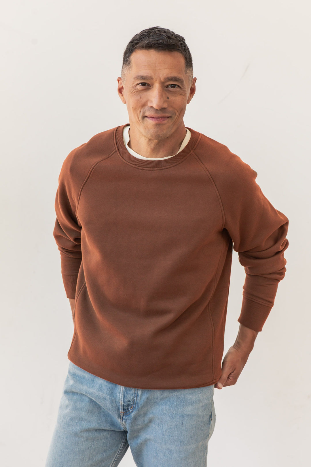 A man with short dark hair wears the Bridge & Burn Lincoln Crew Sweatshirt in Burnt Umber and light blue jeans, standing against a plain white background, smiling softly with his hands behind his back.