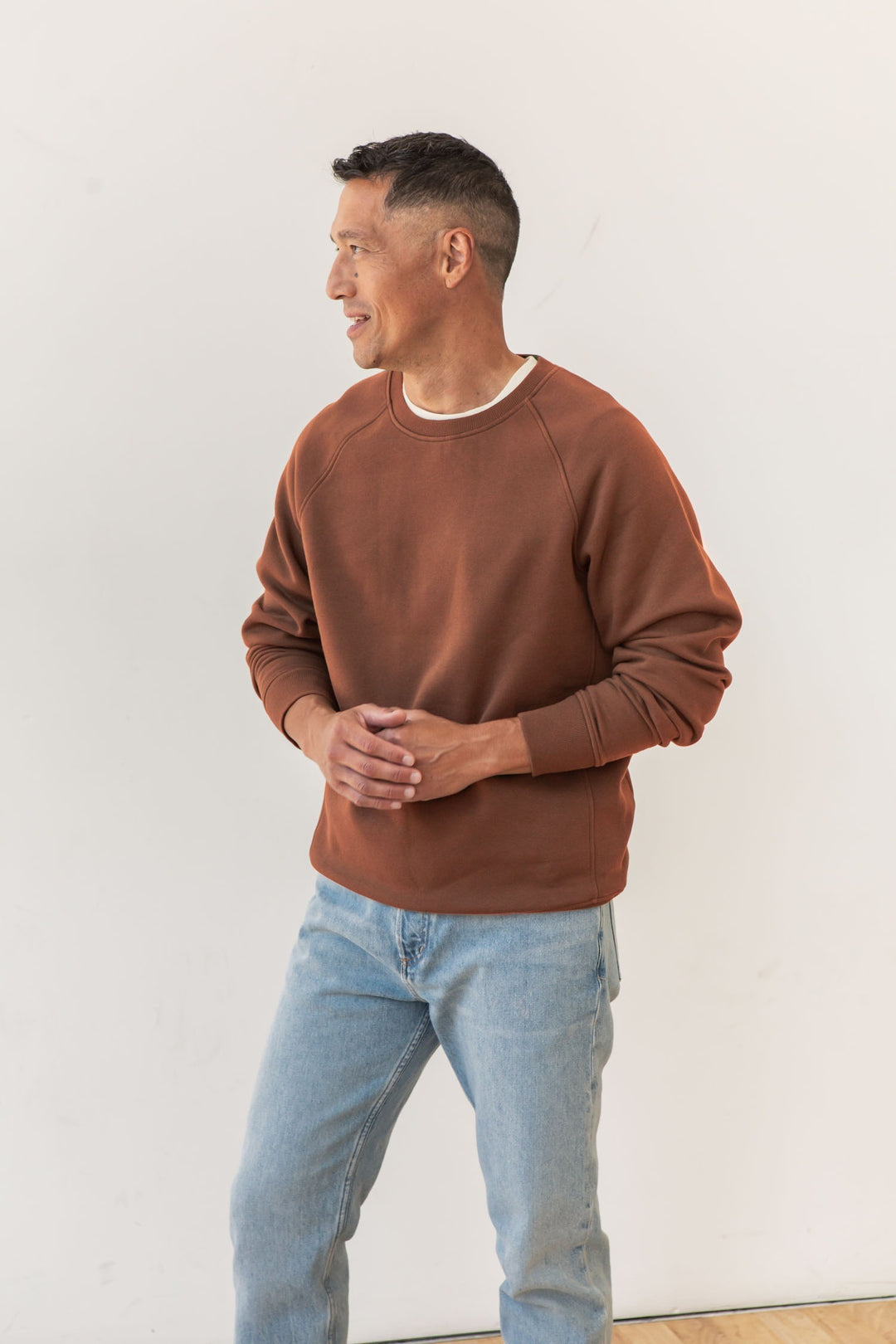 A person with short dark hair wears the Bridge & Burn Lincoln Crew Sweatshirt in Burnt Umber and light blue jeans, standing against a plain light background, looking left and smiling with hands clasped.