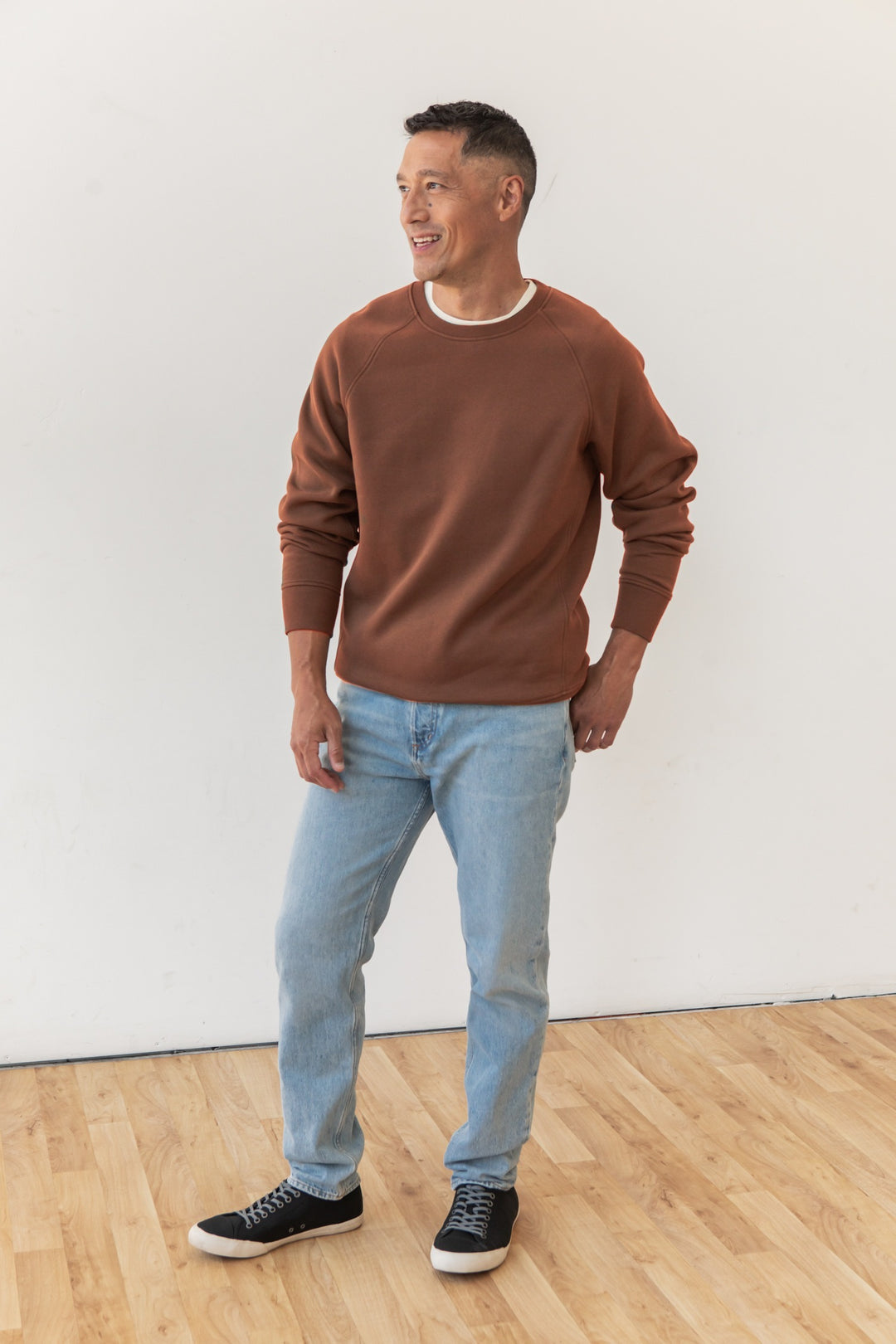 A man stands on a wooden floor against a white wall, wearing Bridge & Burn's Lincoln Crew Sweatshirt in Burnt Umber, light blue jeans, and black sneakers. He looks to the side and smiles.