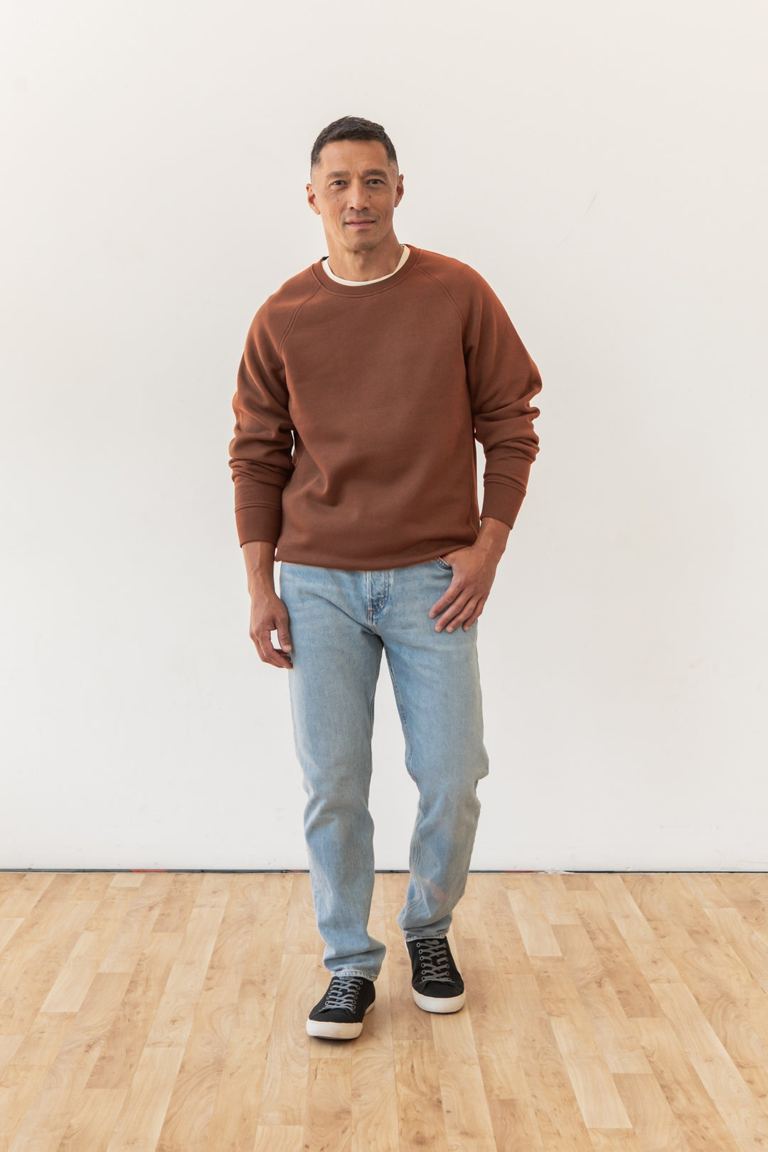 A man stands indoors on a wooden floor against a white wall, wearing the Bridge & Burn Lincoln Crew Sweatshirt in Burnt Umber, light blue jeans, and black sneakers. He looks ahead with a neutral expression and one hand by his side.
