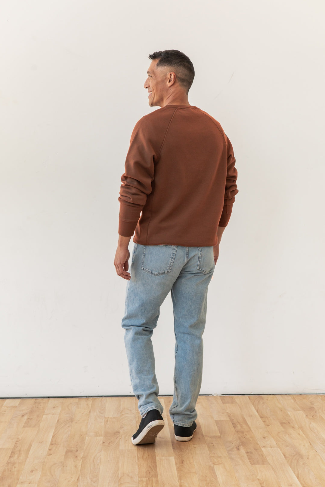 A man with short dark hair wears the Bridge & Burn Lincoln Crew Sweatshirt in Burnt Umber, light blue jeans, and black sneakers. He stands on a wooden floor, facing away from the camera while looking over his left shoulder and smiling.
