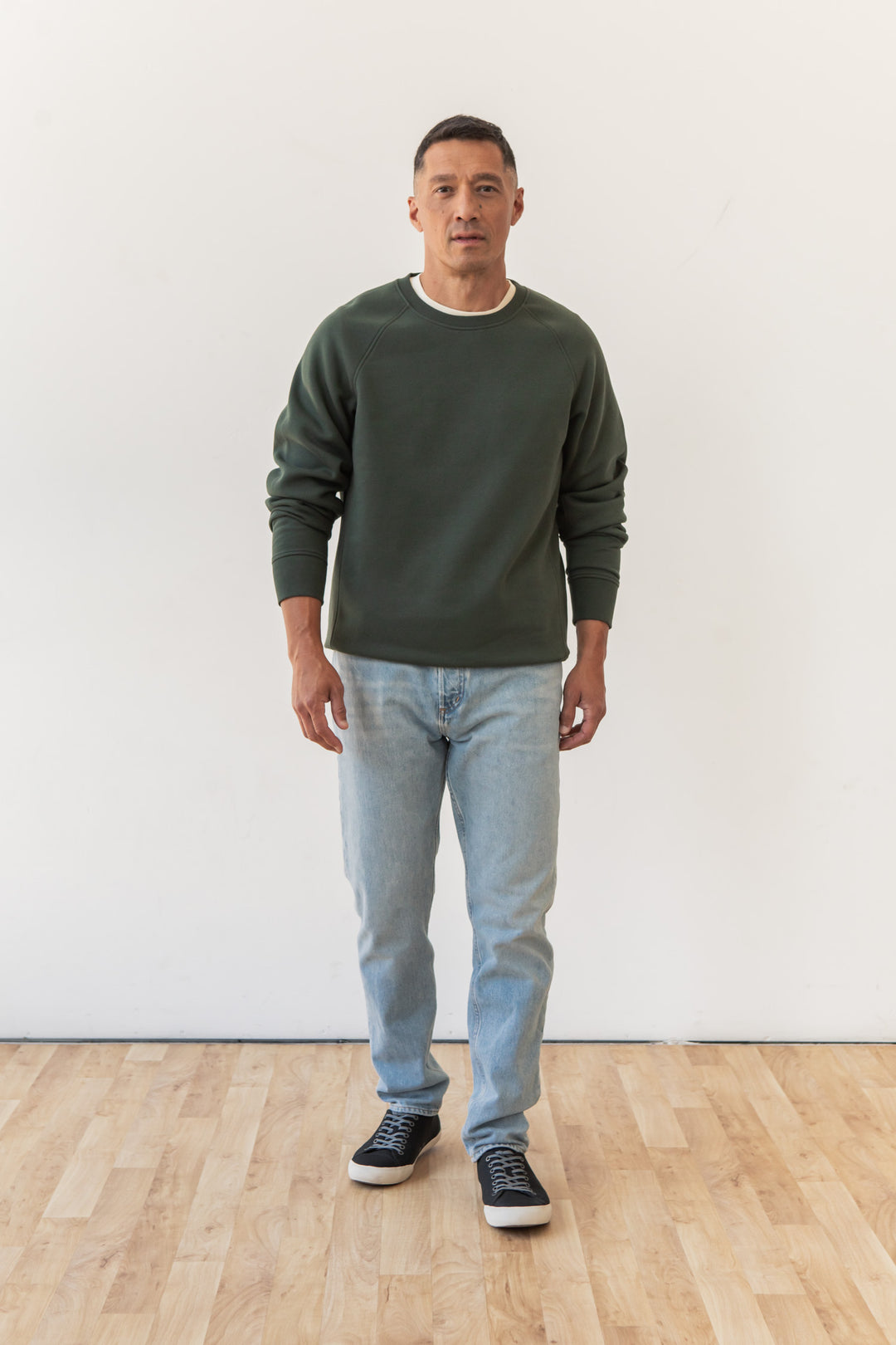 A man stands indoors on a wooden floor by a plain white wall, facing forward and wearing the Bridge & Burn Lincoln Crew Sweatshirt in Forest Night with light blue jeans and black sneakers.