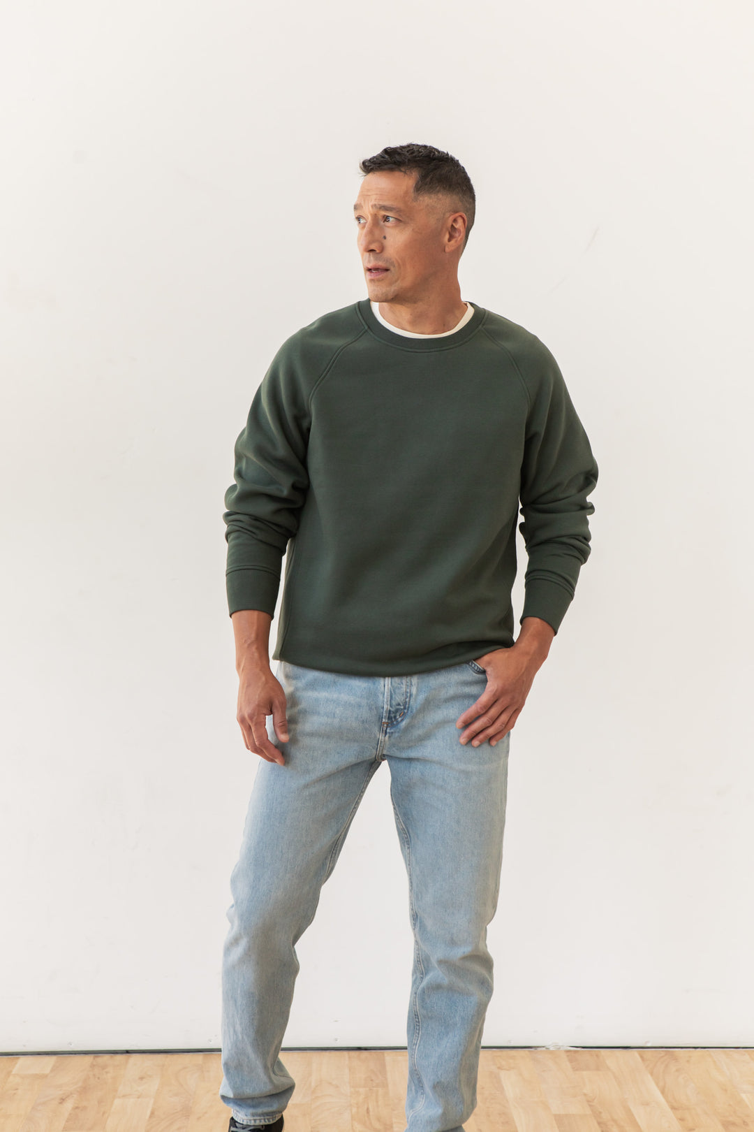 A man stands indoors against a plain white wall, wearing the Bridge & Burn Lincoln Crew Sweatshirt in Forest Night, paired with light blue jeans and sneakers. He looks to the side with one hand in his pocket and the other by his thigh.