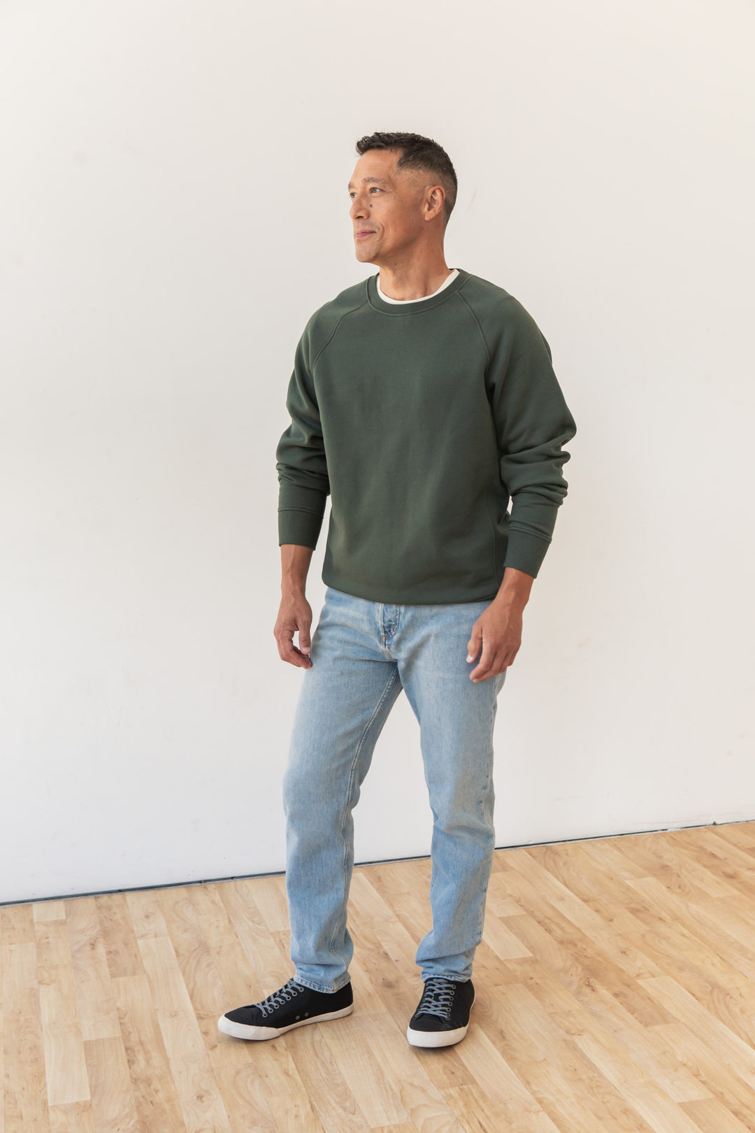 A man stands indoors on a wooden floor, wearing the Bridge & Burn Lincoln Crew Sweatshirt in Forest Night, light blue jeans, and black sneakers. He faces slightly left with a neutral expression against a plain white wall.