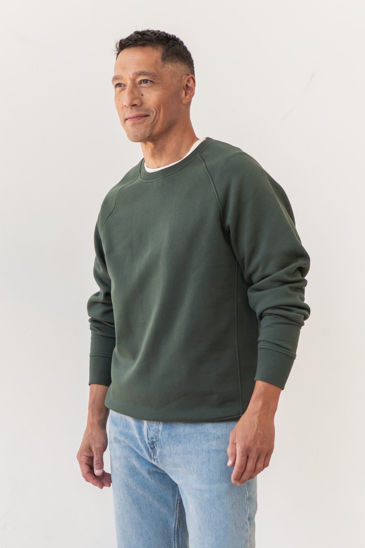 A man stands against a plain white background, wearing the Bridge & Burn Lincoln Crew Sweatshirt in Forest Night and light blue jeans. He looks slightly to the side with a gentle smile.