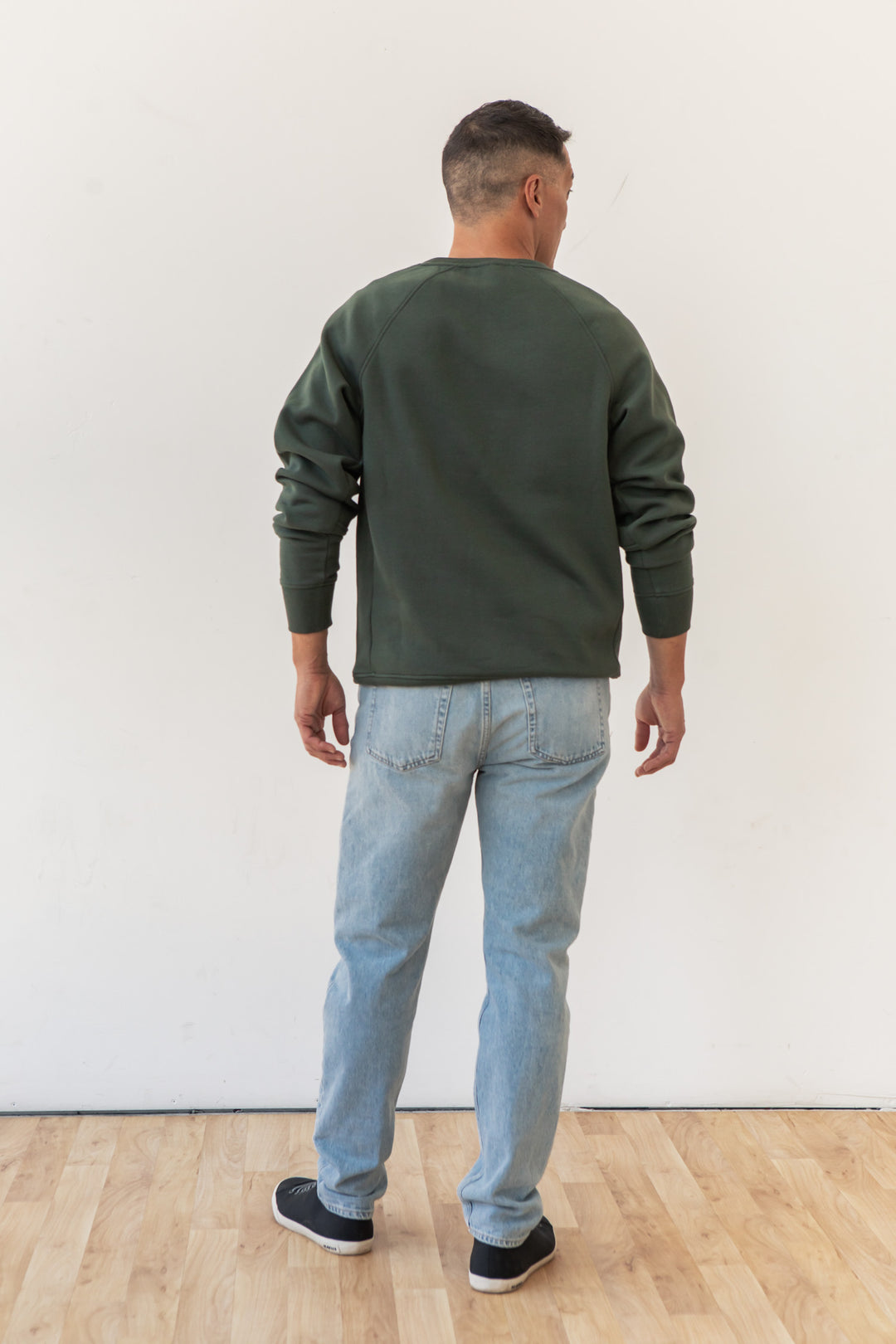 A man stands on a light wood floor, facing away from the camera. He is wearing the Bridge & Burn Lincoln Crew Sweatshirt in Forest Night, paired with light blue jeans and black sneakers against a plain white wall.
