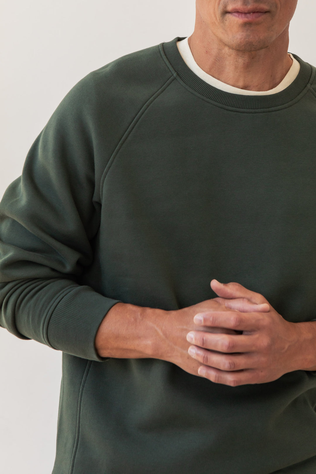 Wearing the Bridge & Burn Lincoln Crew Sweatshirt in Forest Night over a white shirt, a person stands with clasped hands at their chest. Only the lower half of their face and upper body are shown.