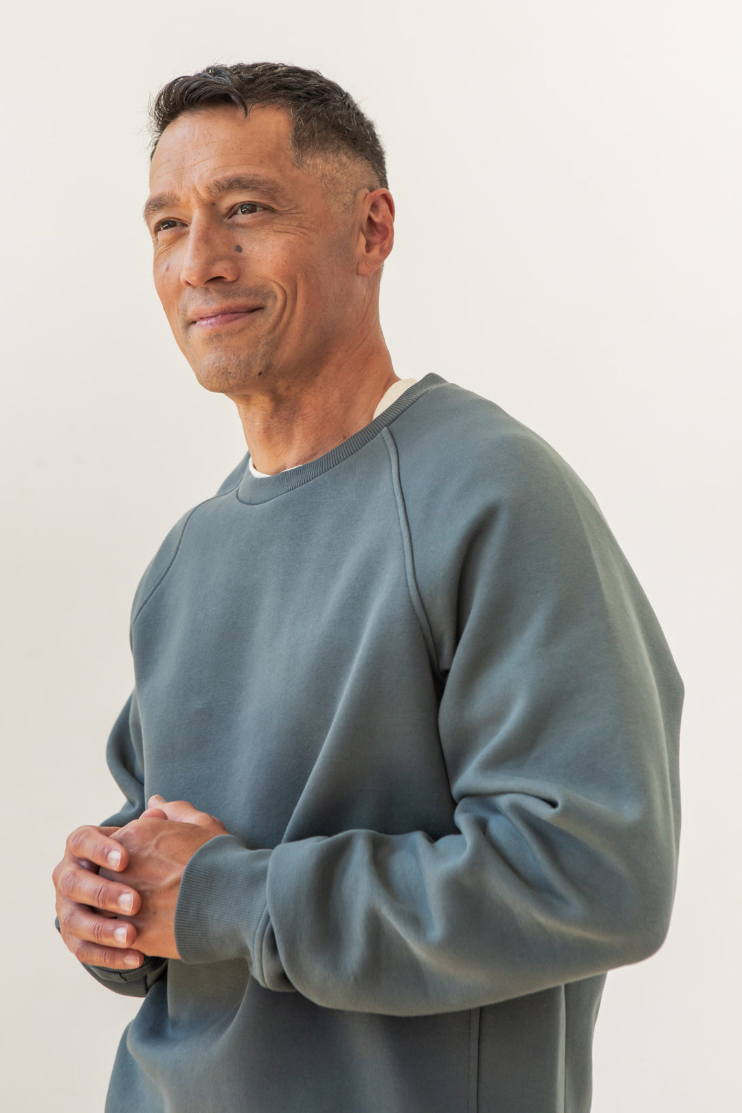 A man wearing the Bridge & Burn Lincoln Crew Sweatshirt in Slate stands against a plain light background, smiling gently with his hands clasped in front of him.