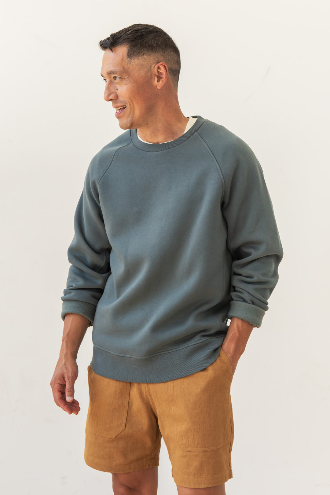 A man with short dark hair wears the Bridge & Burn Lincoln Crew Sweatshirt in Slate, sleeves rolled, paired with light brown shorts. He stands smiling and looking left against a plain white background.