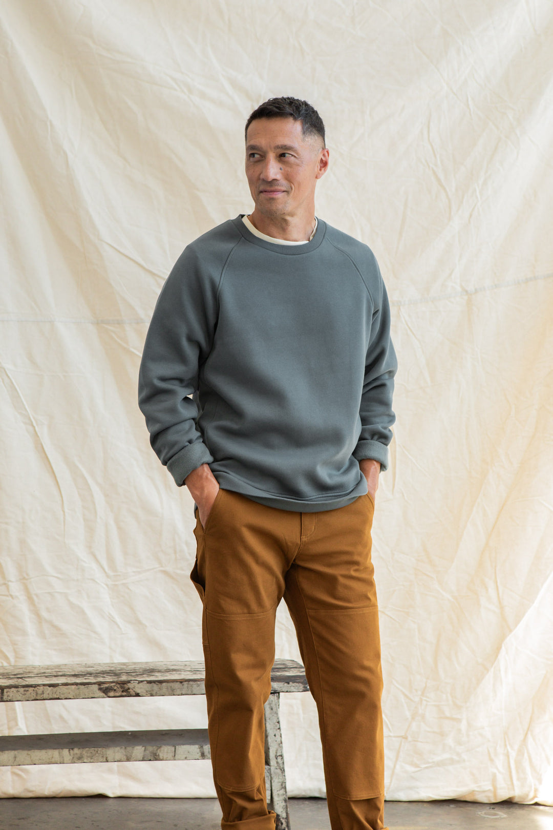 A man with short hair stands before a light fabric backdrop, wearing the Bridge & Burn Lincoln Crew Sweatshirt in Slate and brown pants, hands in pockets, gazing to the side with a soft smile.