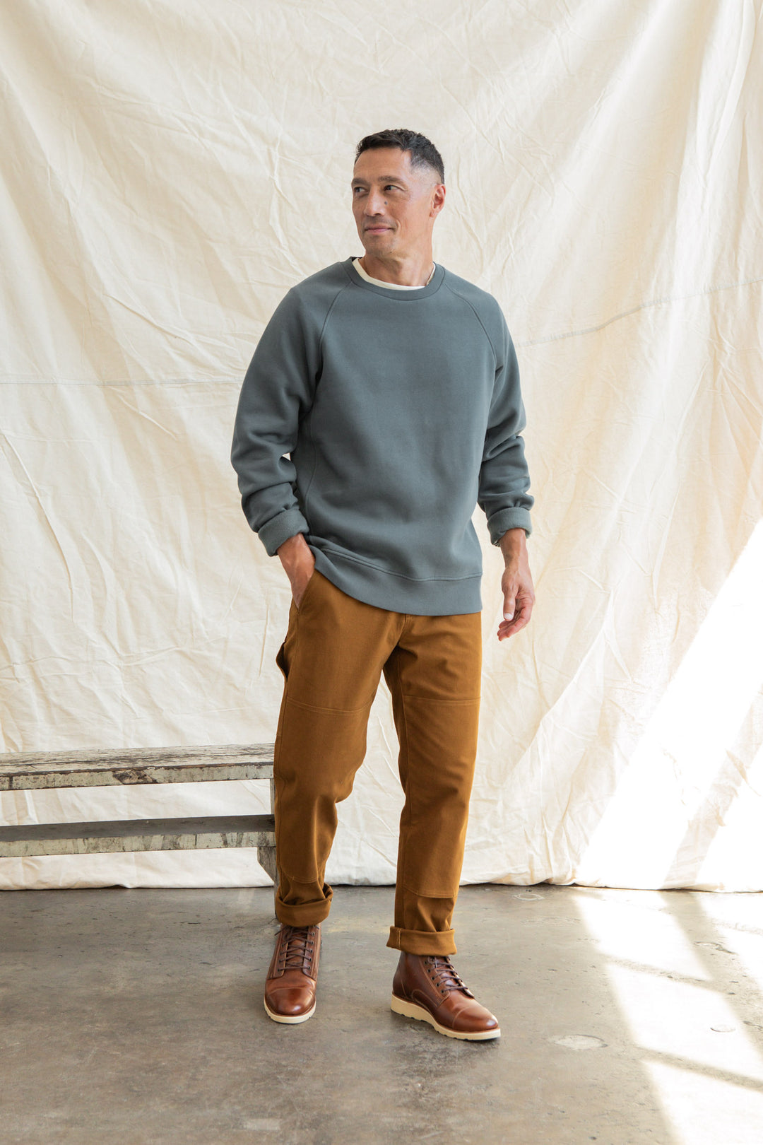 A man stands indoors on a concrete floor, wearing the Bridge & Burn Lincoln Crew Sweatshirt in Slate, brown pants, and brown leather boots. A light-colored fabric backdrop is behind him as he looks slightly to the side with a relaxed posture.