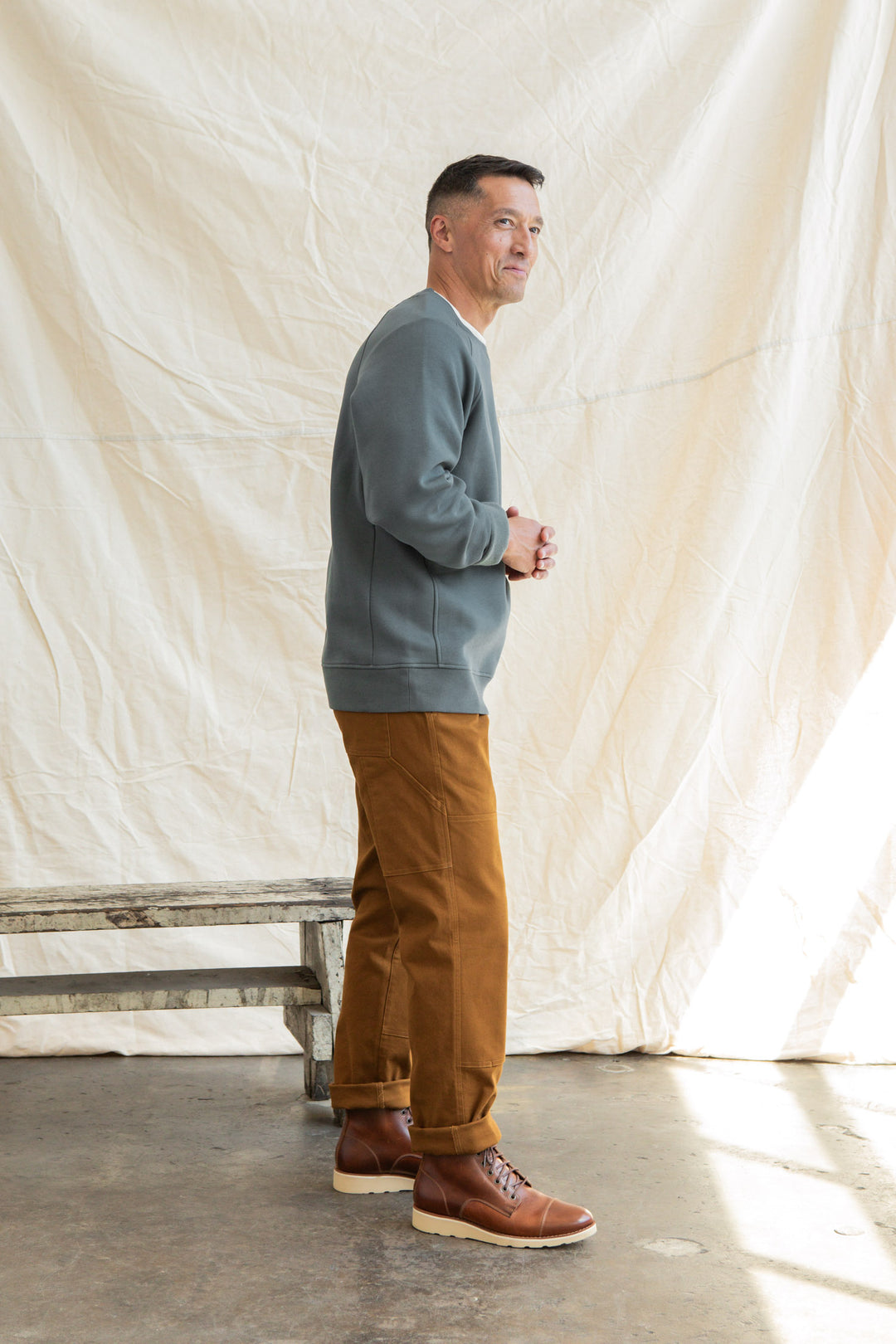 A man stands on a concrete floor, wearing the Bridge & Burn Lincoln Crew Sweatshirt in Slate, brown pants, and brown boots. He faces sideways with hands clasped beside a rustic wooden bench and a cream fabric backdrop.