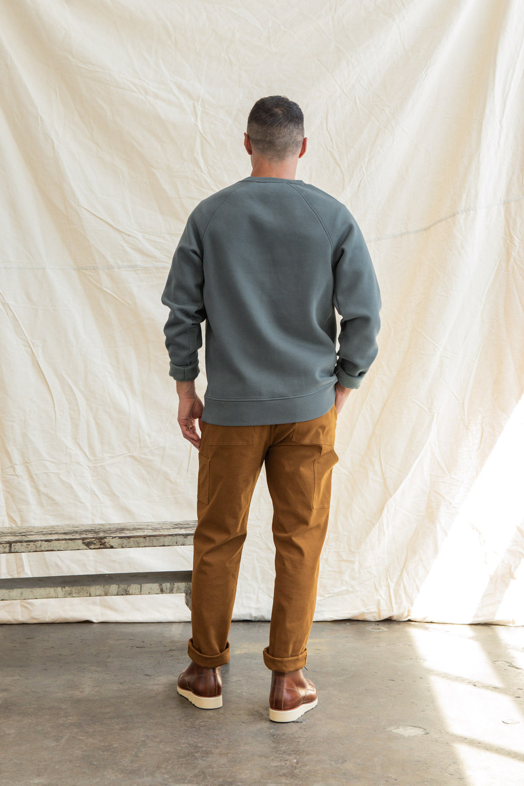 A man stands indoors facing a cream-colored fabric backdrop, wearing the Bridge & Burn Lincoln Crew Sweatshirt in Slate with brown pants and shoes. Photographed from behind, his hands rest at his sides.