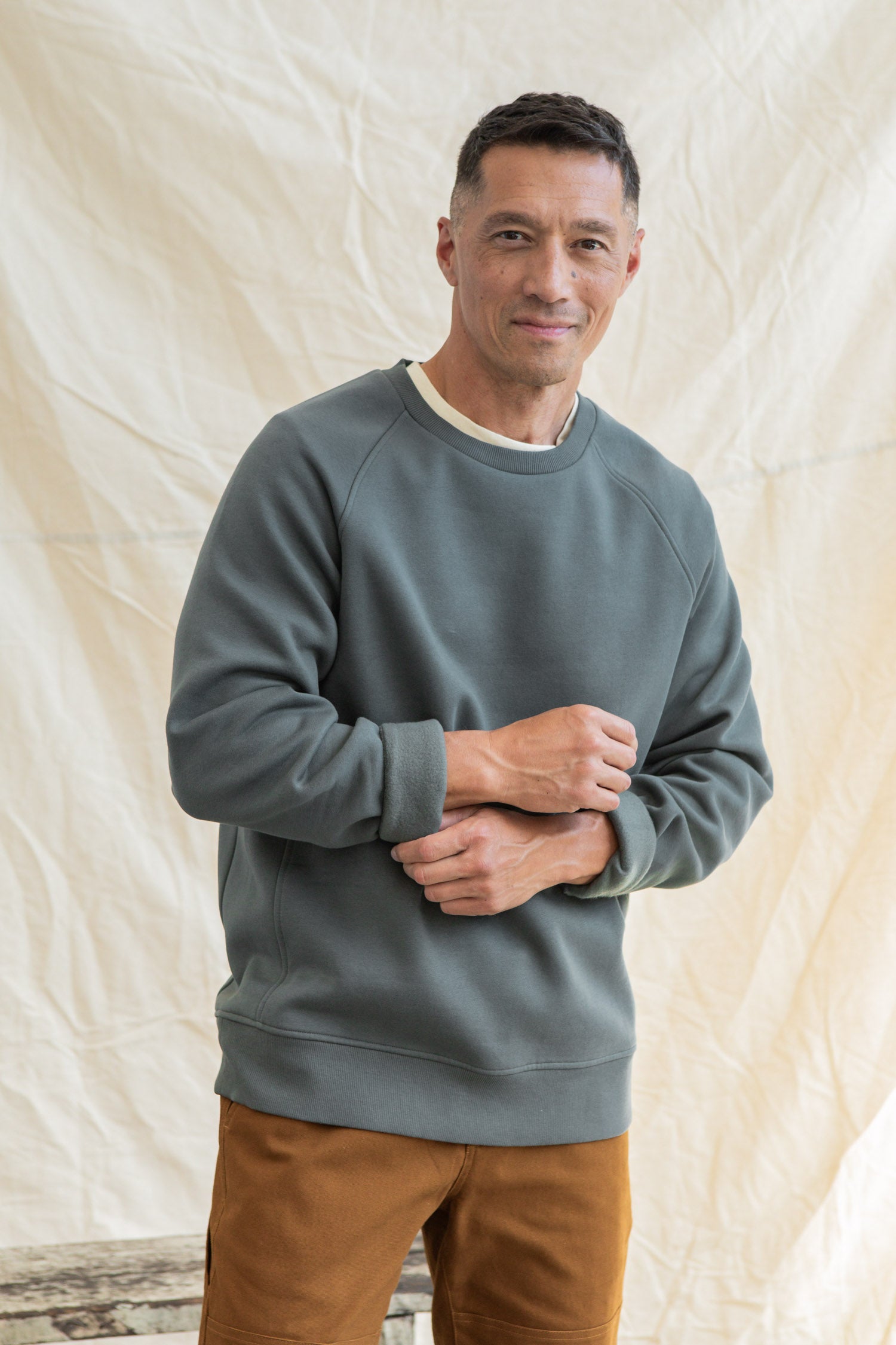 A man with short dark hair smiles gently at the camera, wearing the Bridge & Burn Lincoln Crew Sweatshirt in Slate over a white shirt and brown pants, standing before a light-colored fabric backdrop.