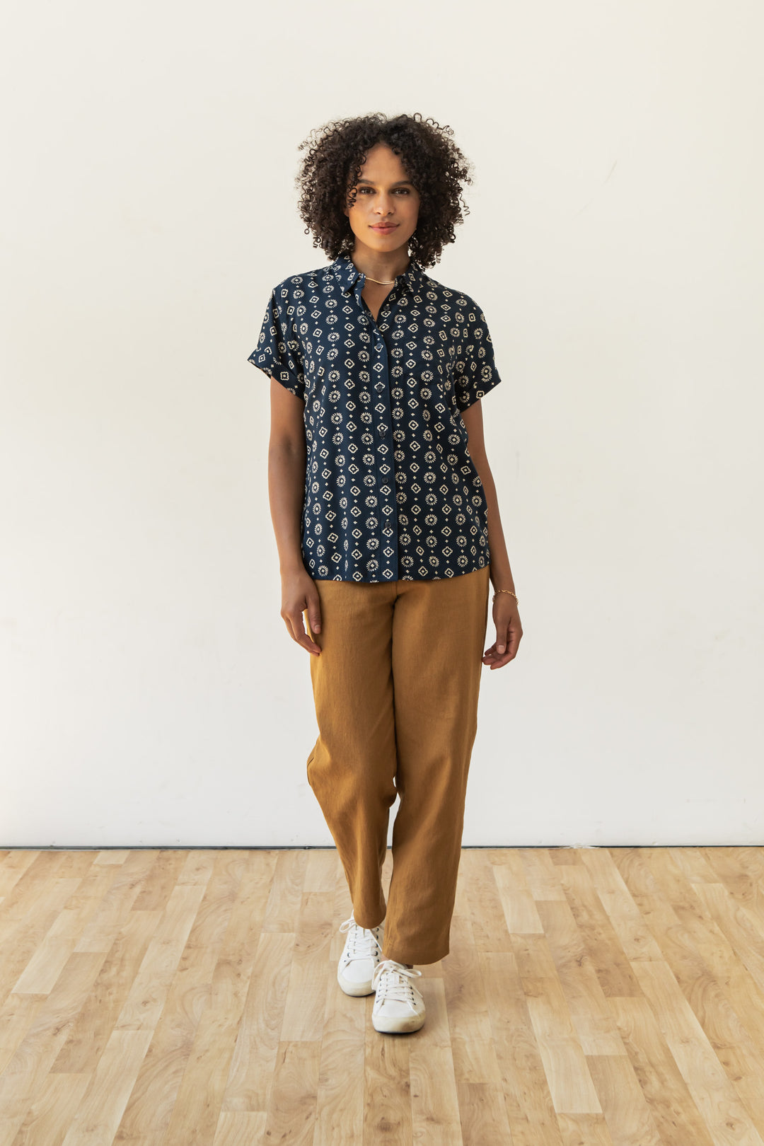 A person with curly hair wears the Bridge & Burn Bea Shirt in Midnight Sundance, tan pants, and white sneakers while standing on a light wooden floor against a plain white wall.