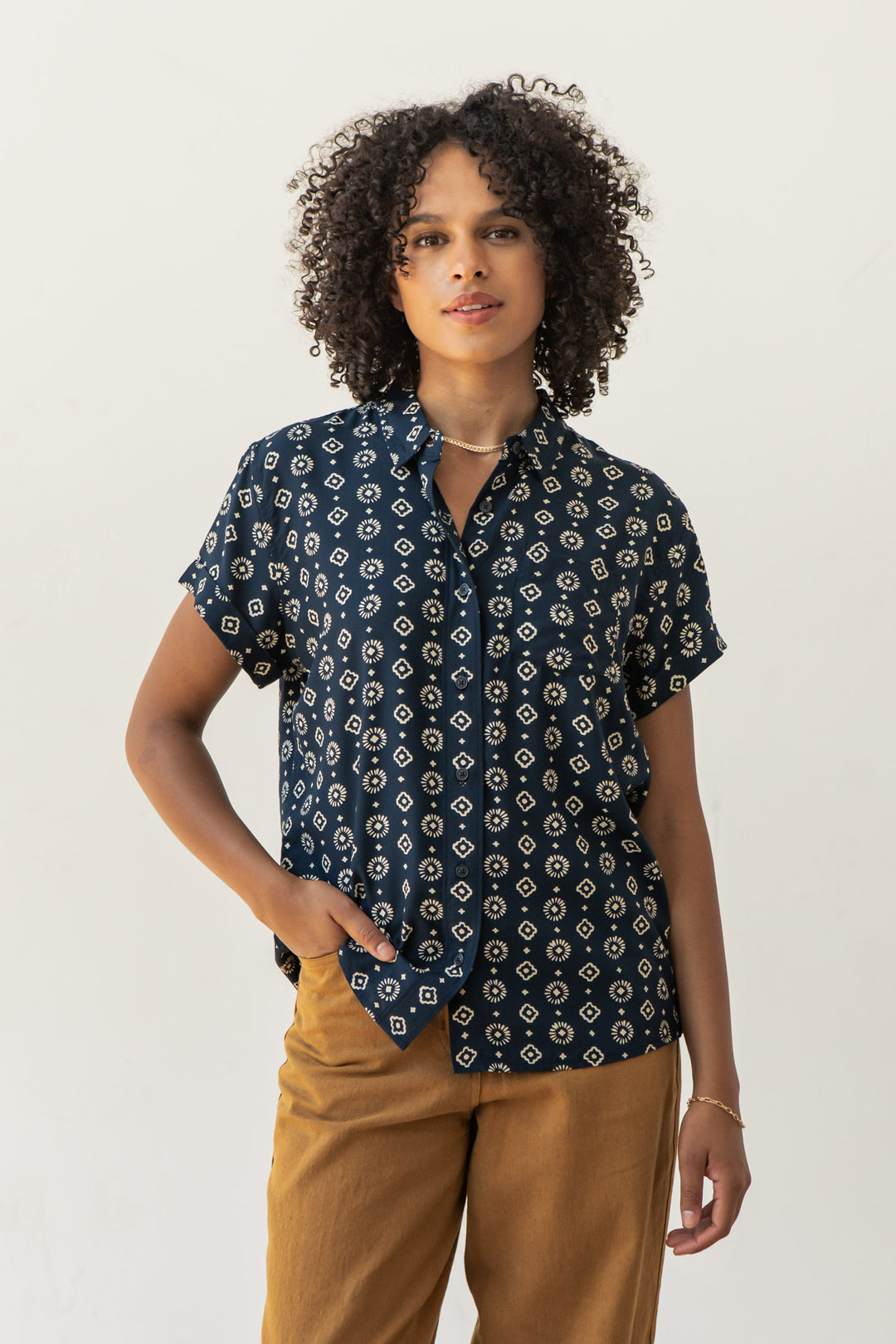 A person with curly hair wears the Bea Shirt in Midnight Sundance by Bridge & Burn and tan pants, standing against a plain light background while looking at the camera with one hand in their pocket.