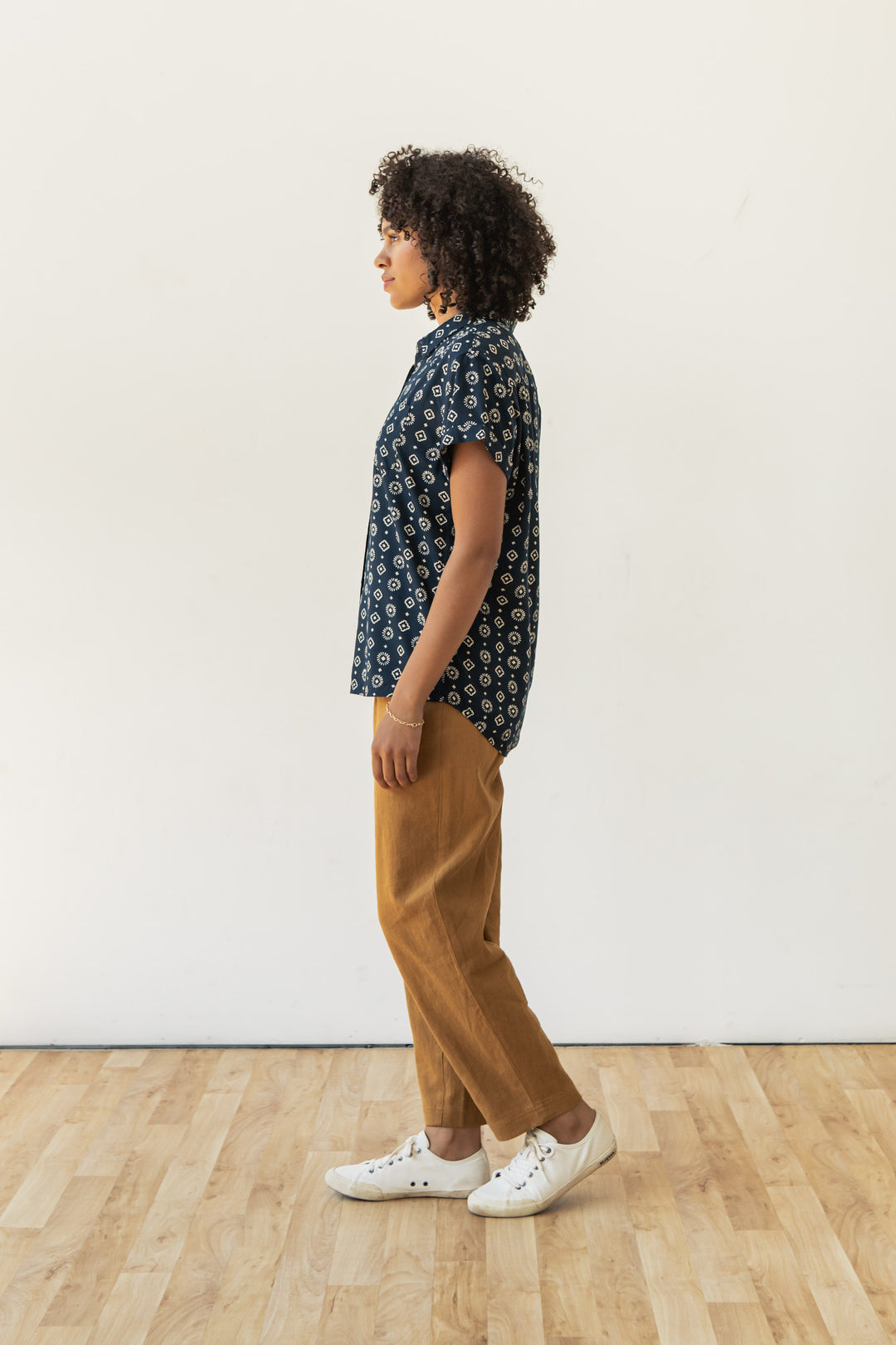 A person with curly hair stands in profile on a wooden floor, wearing the Bridge & Burn Bea Shirt in Midnight Sundance, tan pants, and white sneakers against a plain light-colored wall.