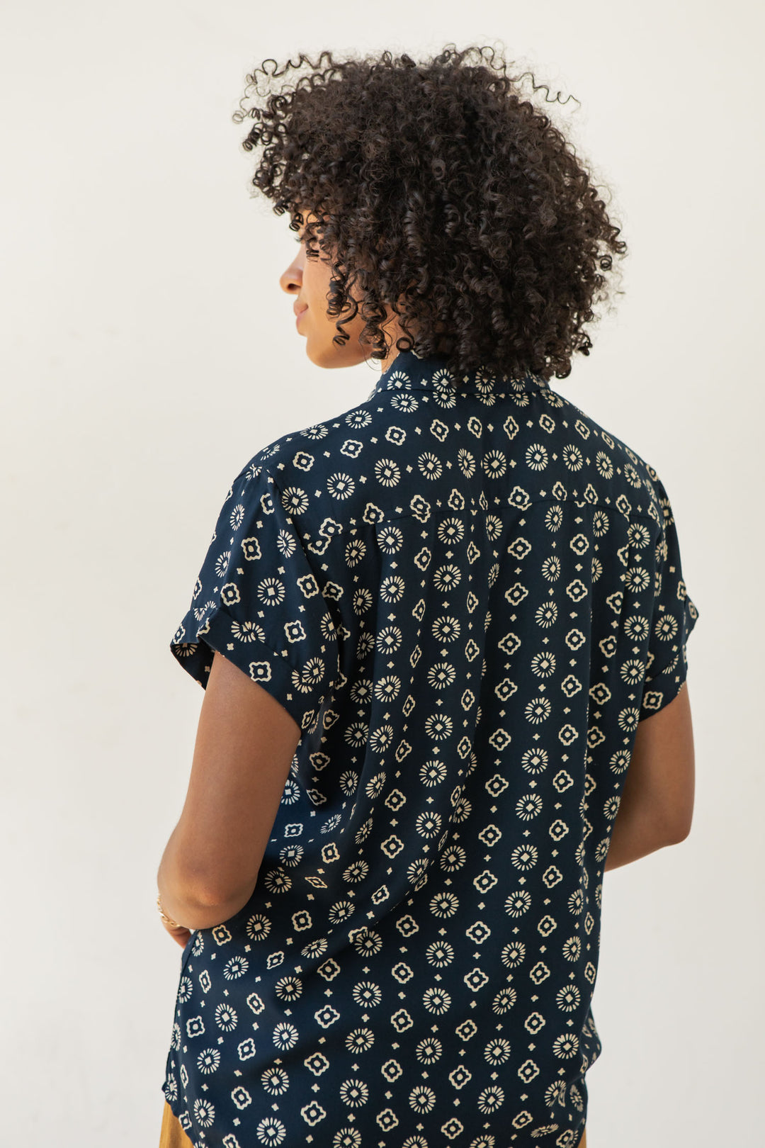A person with curly hair wears the Bridge & Burn Bea Shirt in Midnight Sundance, a tailored-fit navy short-sleeve button-down with a white geometric pattern, standing facing away from the camera against a plain light background.
