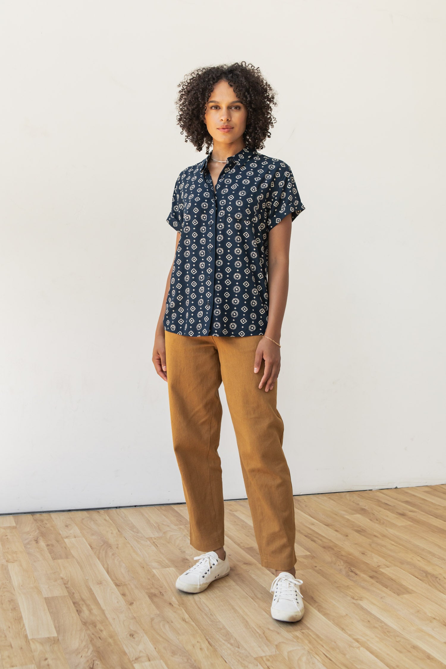 A person with curly hair stands on a wooden floor against a plain white wall, wearing the Bridge & Burn Bea Shirt in Midnight Sundance, paired with light brown pants and white sneakers.