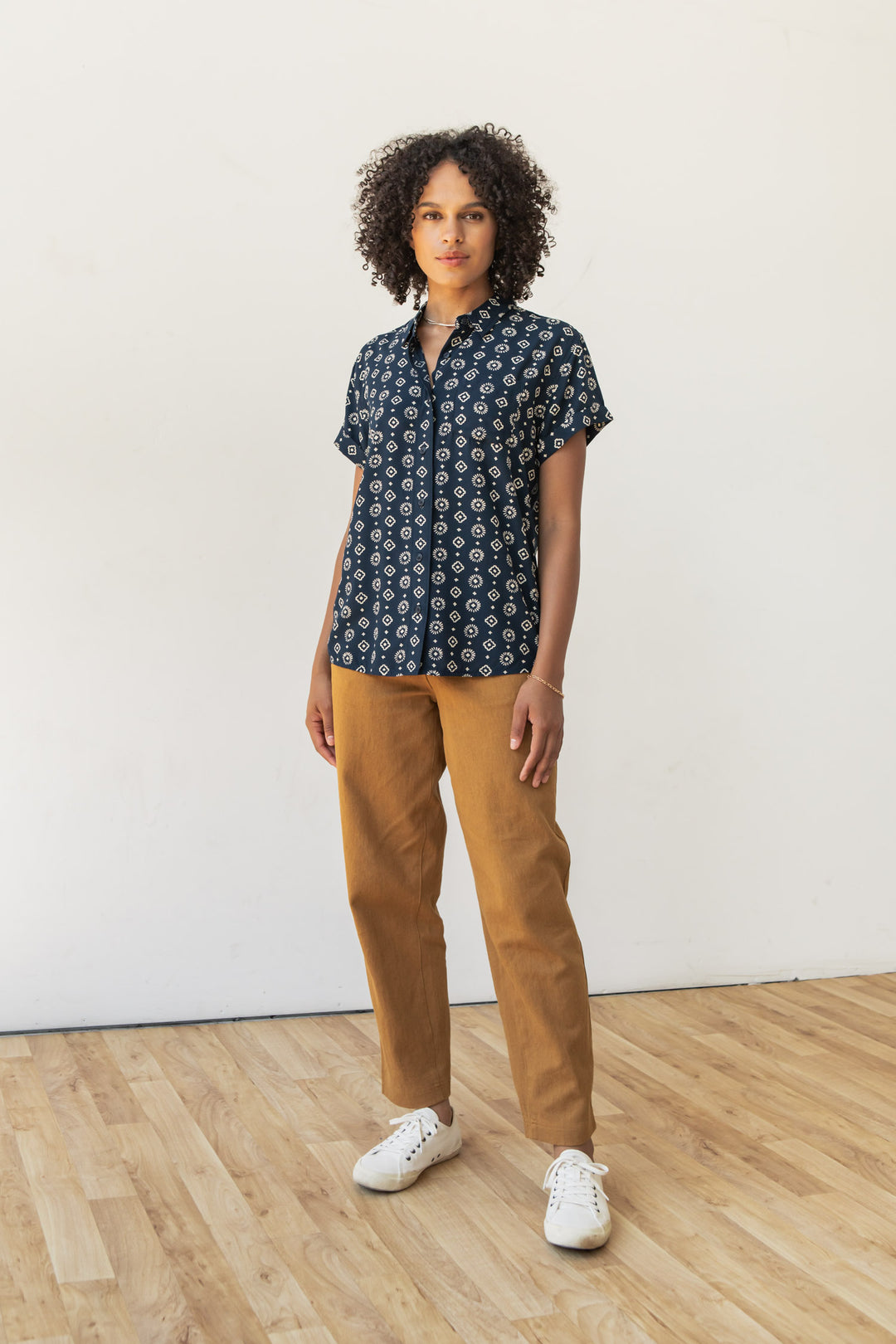 A person with curly hair stands on a wooden floor against a plain white wall, wearing the Bridge & Burn Bea Shirt in Midnight Sundance, paired with light brown pants and white sneakers.