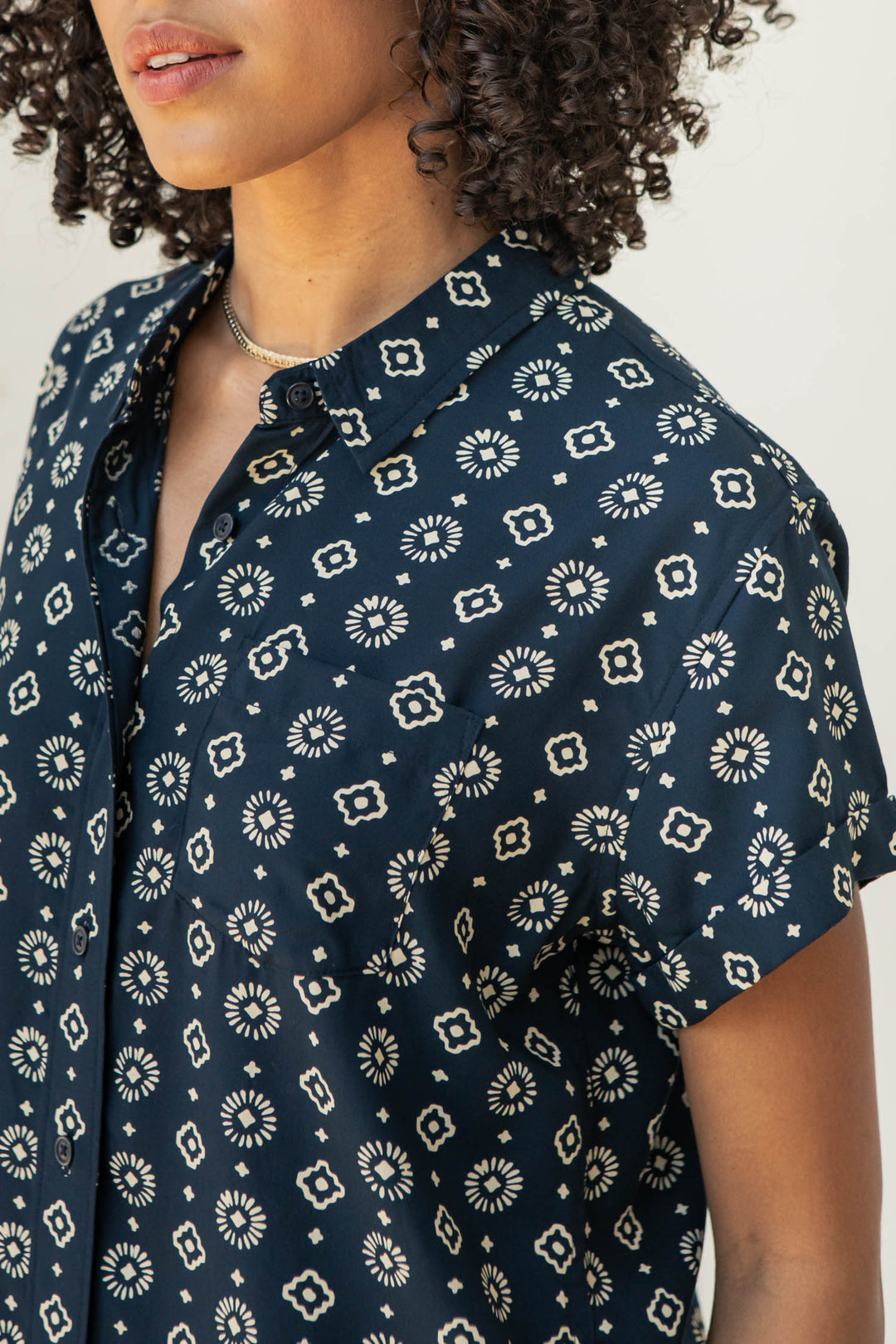 A woman with curly hair models the Bridge & Burn Bea Shirt in Midnight Sundance, a tailored-fit navy short-sleeve button-down with a cream geometric floral pattern. Only her lower face and torso are shown against a light background.