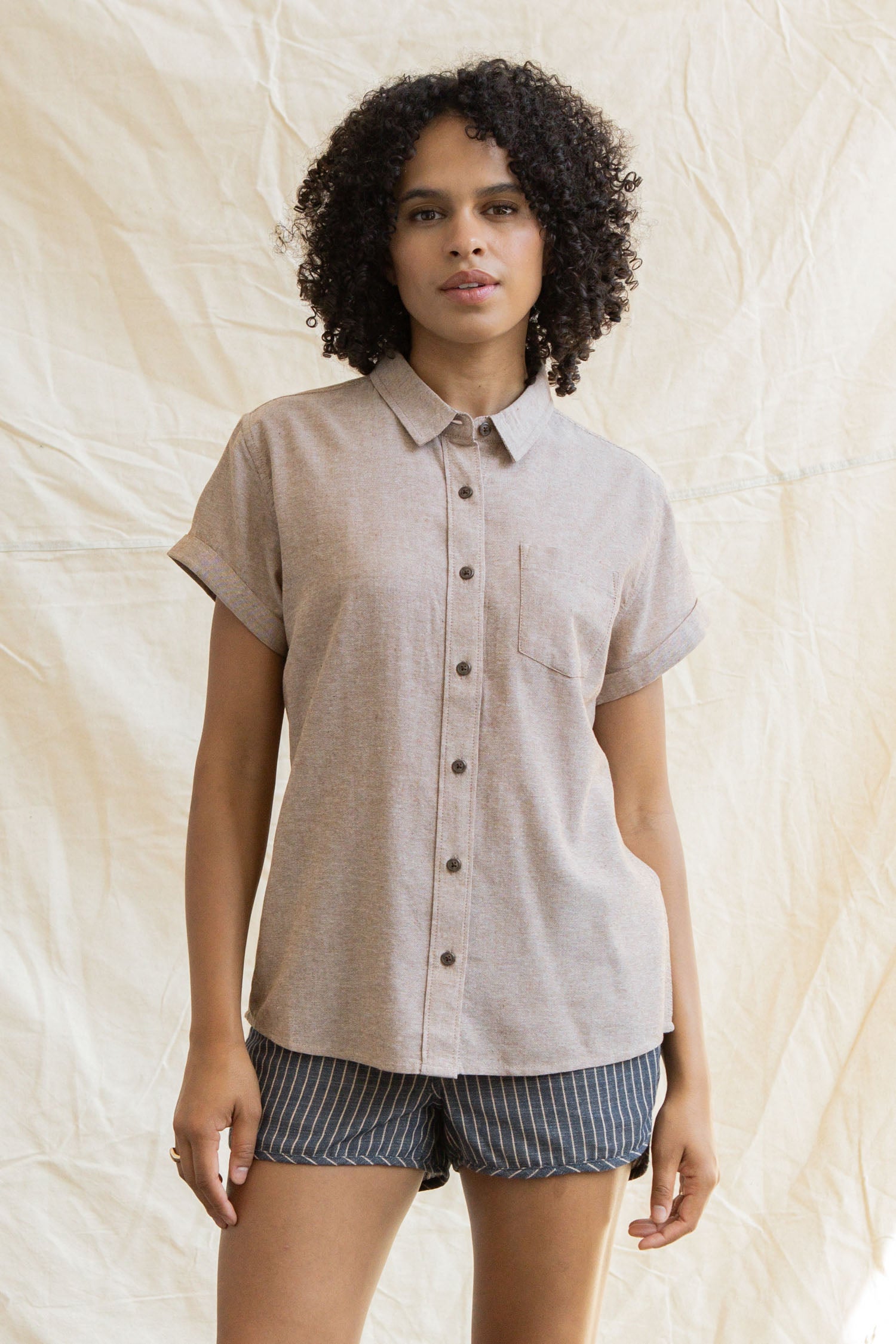 A person with curly hair wears the Bridge & Burn Bea Shirt in Rust Chambray and stands in front of a light fabric backdrop.
