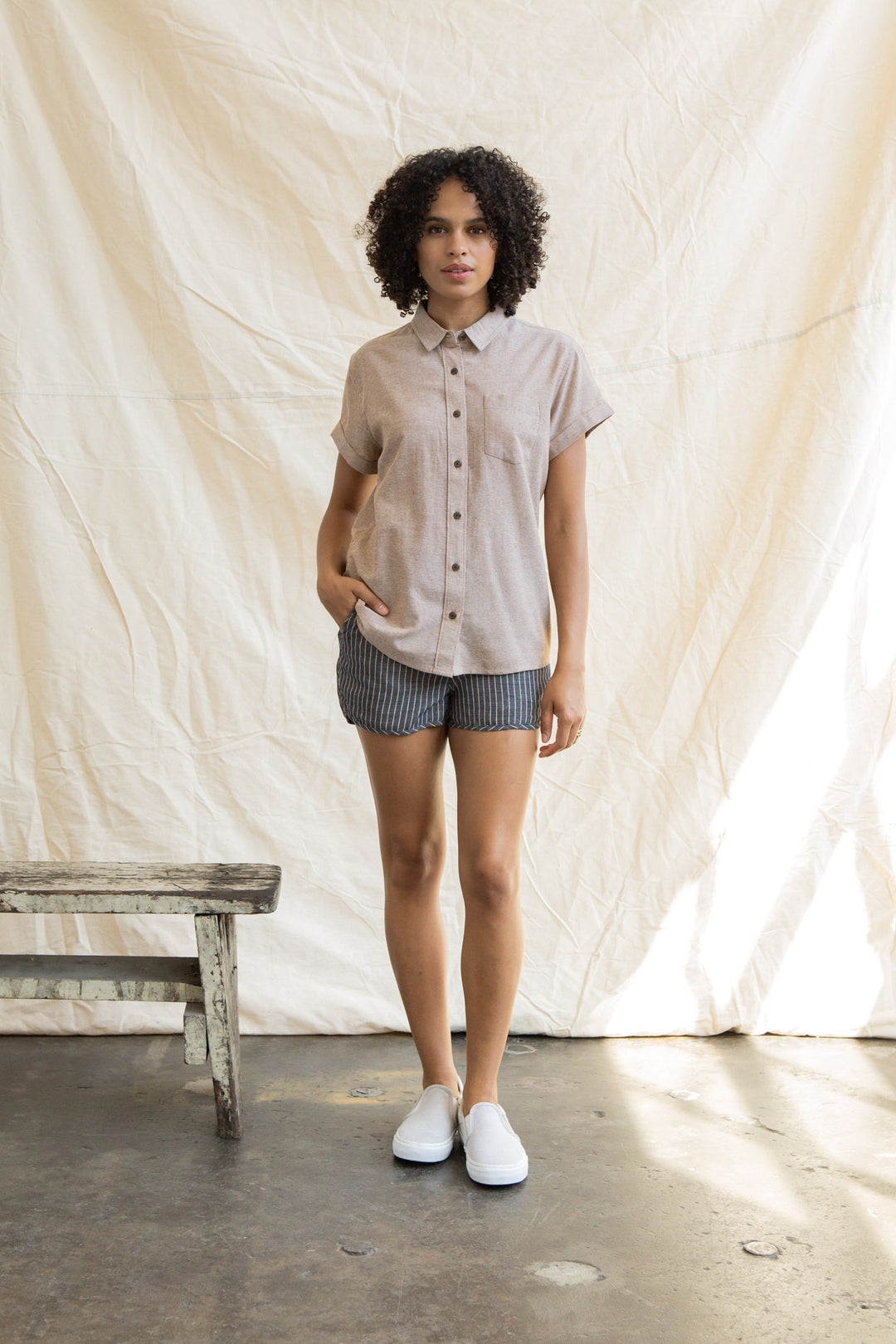 A woman with curly hair stands indoors by a beige fabric backdrop, wearing the Bridge & Burn Bea Shirt in Rust Chambray, striped shorts, and white slip-on shoes. One hand is in her pocket and a rustic wooden bench is next to her.