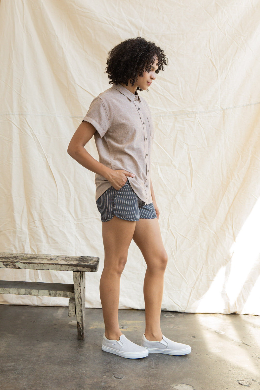 A person with curly hair stands sideways, wearing the Bridge & Burn Bea Shirt in Rust Chambray, striped shorts, and slip-on shoes beside a rustic wooden bench against a light fabric backdrop.
