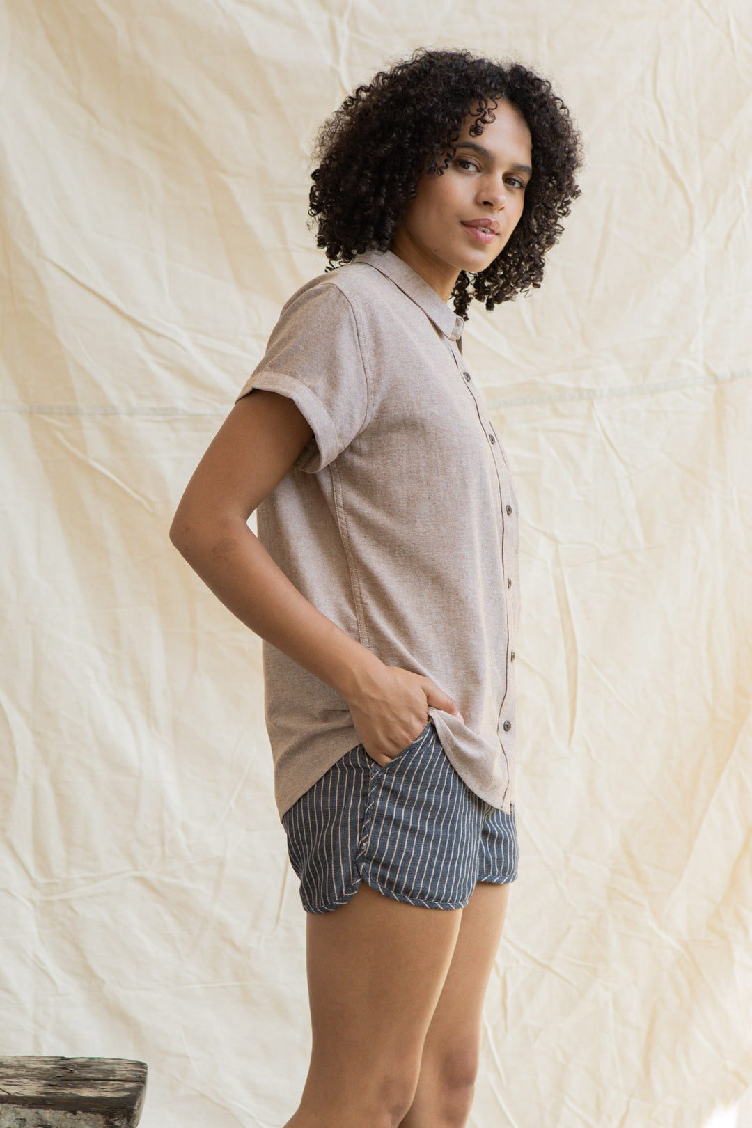 A person with curly hair stands sideways, facing the camera, wearing the Bridge & Burn Bea Shirt in Rust Chambray and blue pinstriped shorts. One hand is in their pocket as they pose against a light fabric background.