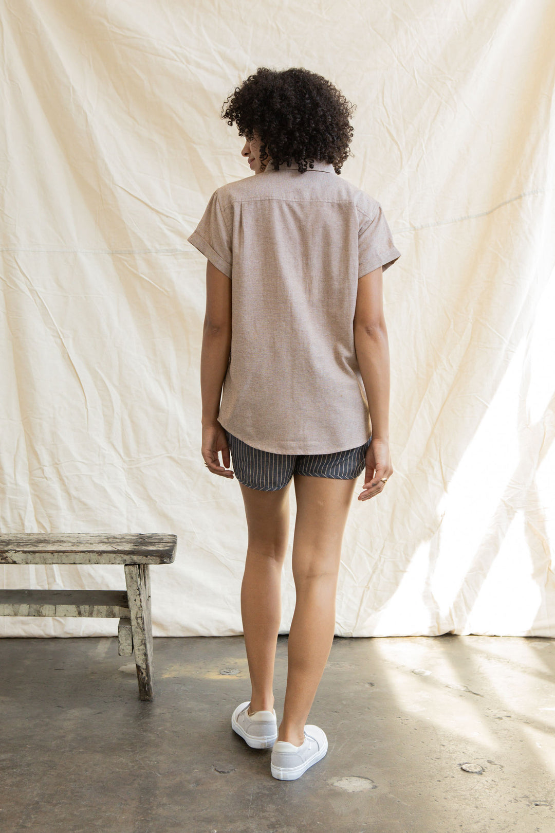 A person with curly hair stands indoors facing away, wearing the Bridge & Burn Bea Shirt in Rust Chambray. They're near a rustic wooden bench and a cream-colored backdrop as sunlight streams in from the side.