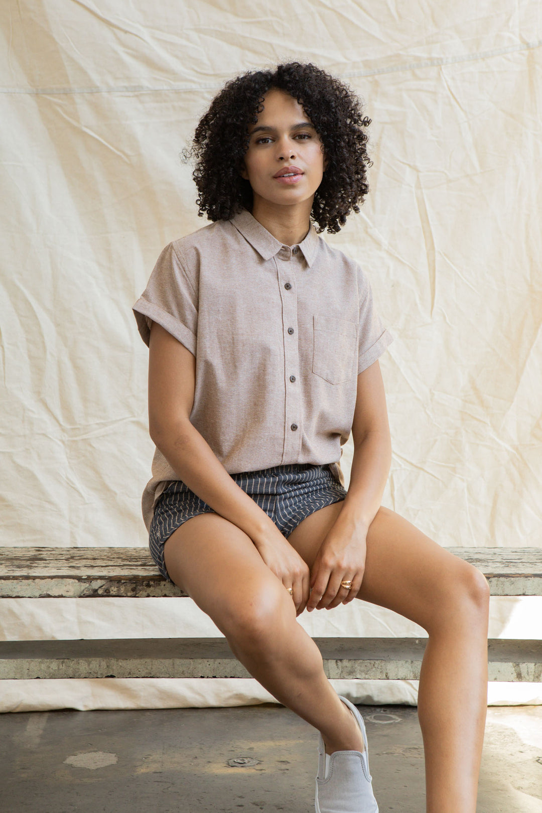 A woman with curly hair wears the Bridge & Burn Bea Shirt in Rust Chambray and patterned shorts, sitting on a wooden bench against a light fabric backdrop. She looks at the camera with a relaxed expression.