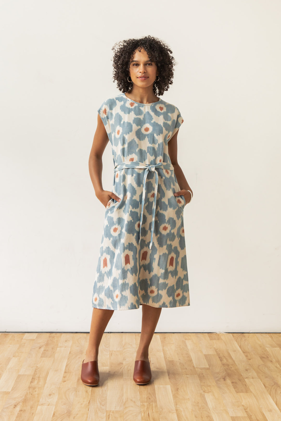 A person with curly hair stands on a wooden floor, wearing the Bridge & Burn Eloise Dress in Polar Blossom—a blue, 100% cotton A-line dress with short sleeves and waist belt—with brown slip-ons, hands in pockets, facing forward against a plain background.