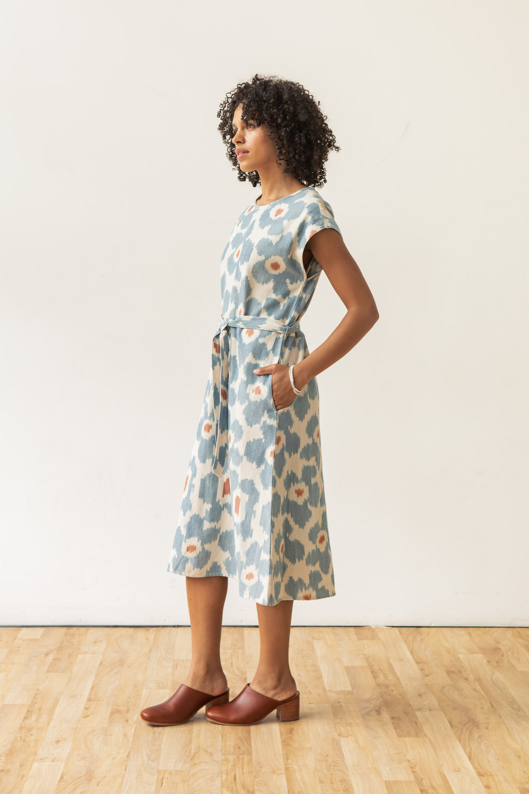 A person stands on a wooden floor against a plain white background, wearing the Bridge & Burn Eloise Dress in Polar Blossom—a light blue, white, and orange 100% cotton A-line dress with short sleeves and a belt—paired with brown clogs.