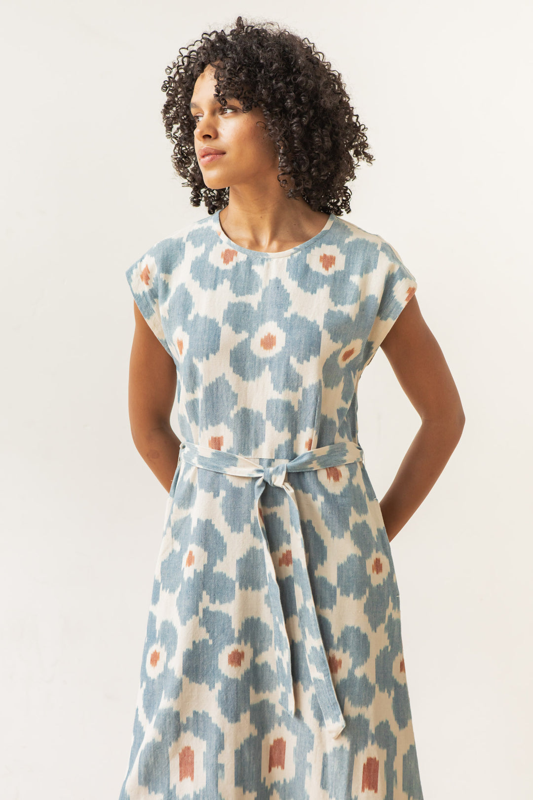 A woman with curly hair wears the Bridge & Burn Eloise Dress in Polar Blossom, a short-sleeved, belted A-line dress with abstract blue and white floral patterns and orange accents, standing against a plain light background, looking left.