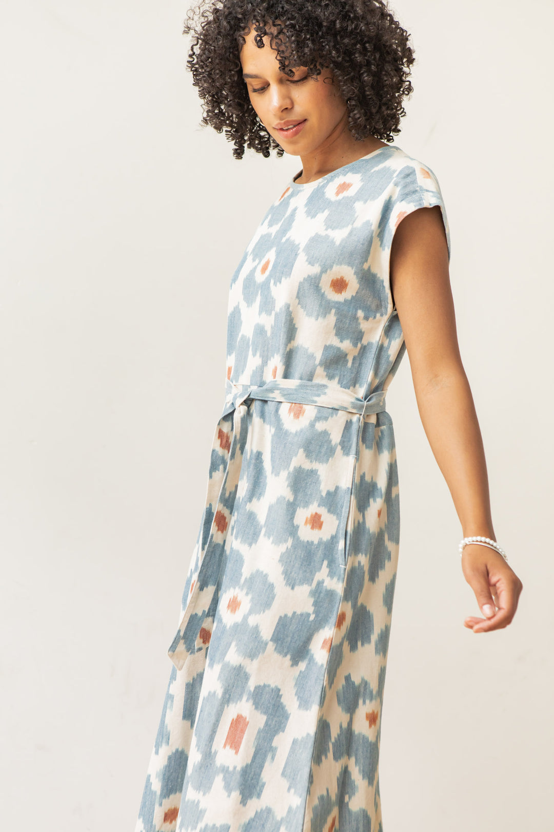 A woman with curly hair wears the Bridge & Burn Eloise Dress in Polar Blossom—an A-line design featuring a blue and orange abstract floral print—standing against a plain light background, gazing slightly downward.