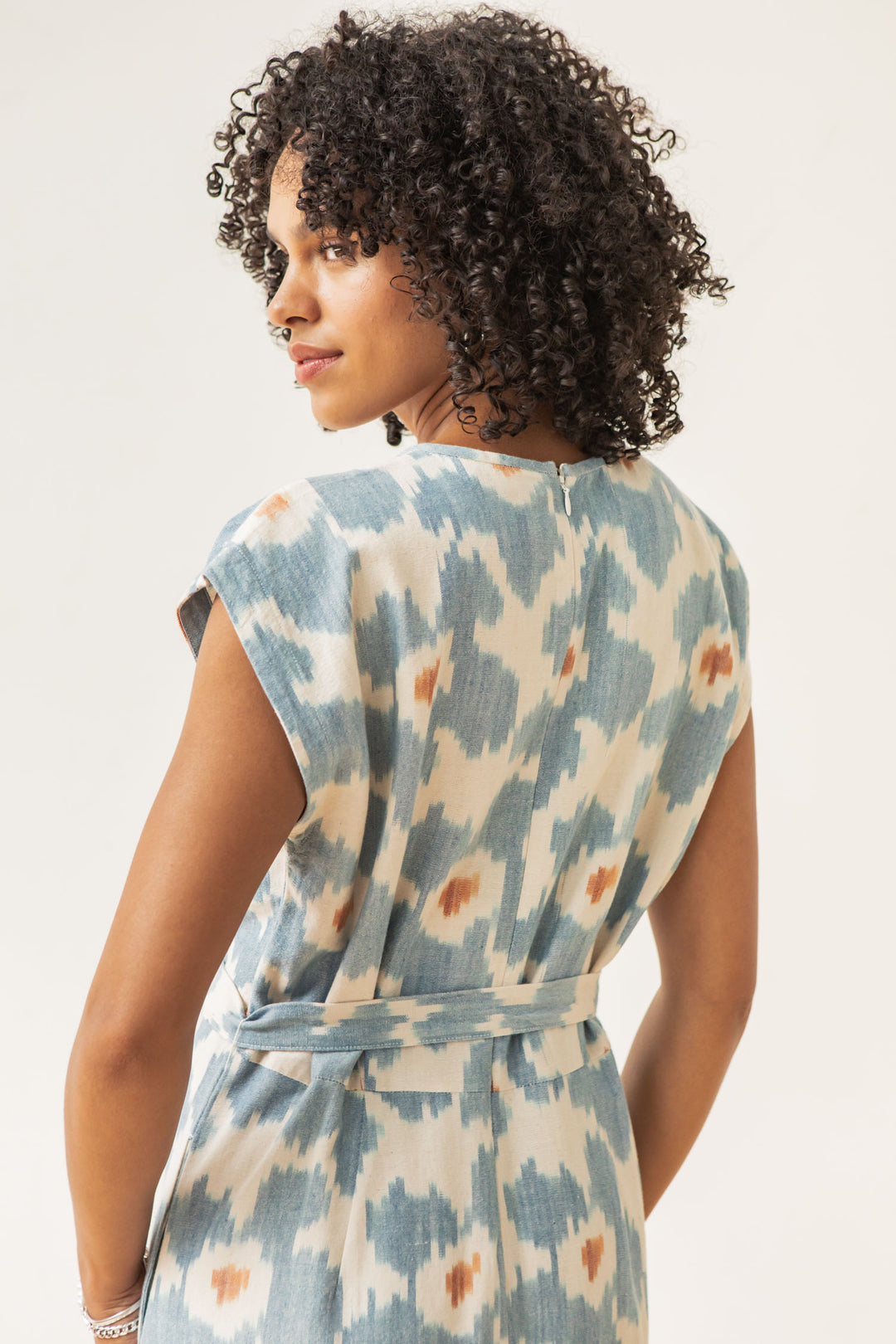 A woman with curly hair models the Bridge & Burn Eloise Dress in Polar Blossom, a sleeveless, belted 100% cotton dress with a blue and beige abstract print. She poses back to the camera, glancing over her shoulder against a light backdrop.
