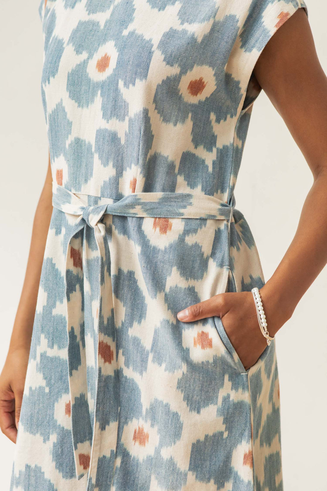 A person wearing the Eloise Dress in Polar Blossom by Bridge & Burn, a sleeveless blue and white A-line dress with a tie belt, one hand in the pocket, accessorized with a bracelet.