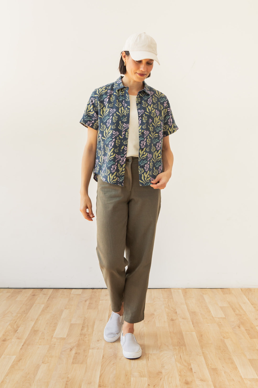 A person stands indoors on a wooden floor, wearing a white cap and shirt, the Bridge & Burn Greer Cropped Shirt in Spring Kelp (boxy fit, botanical print), olive-green pants, and white slip-on shoes.