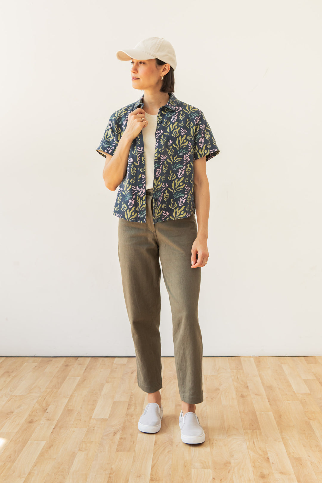 A person stands on a light wooden floor against a plain white wall, wearing the Bridge & Burn Greer Cropped Shirt in Spring Kelp over a white top, with olive green pants, a white cap, and white slip-on shoes.