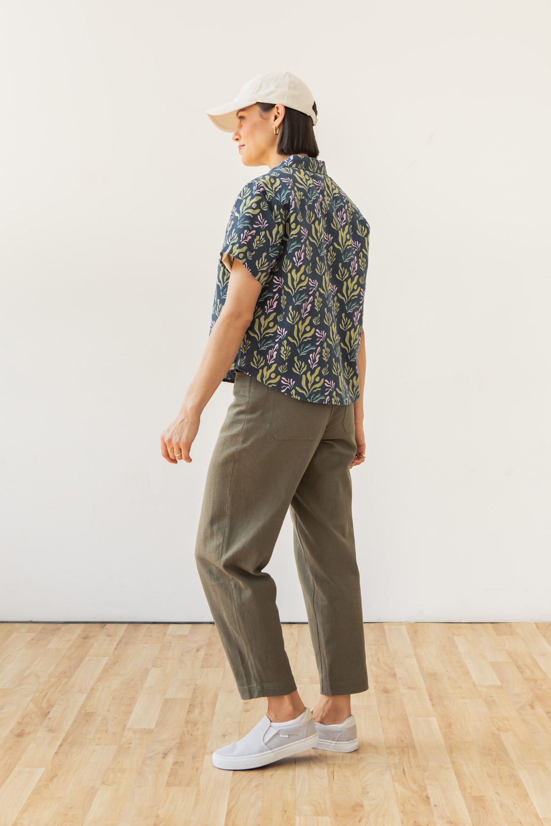 A person stands sideways on a wooden floor, wearing the Bridge & Burn Greer Cropped Shirt in Spring Kelp, with a white cap, olive green pants, and white slip-on shoes against a plain white wall.