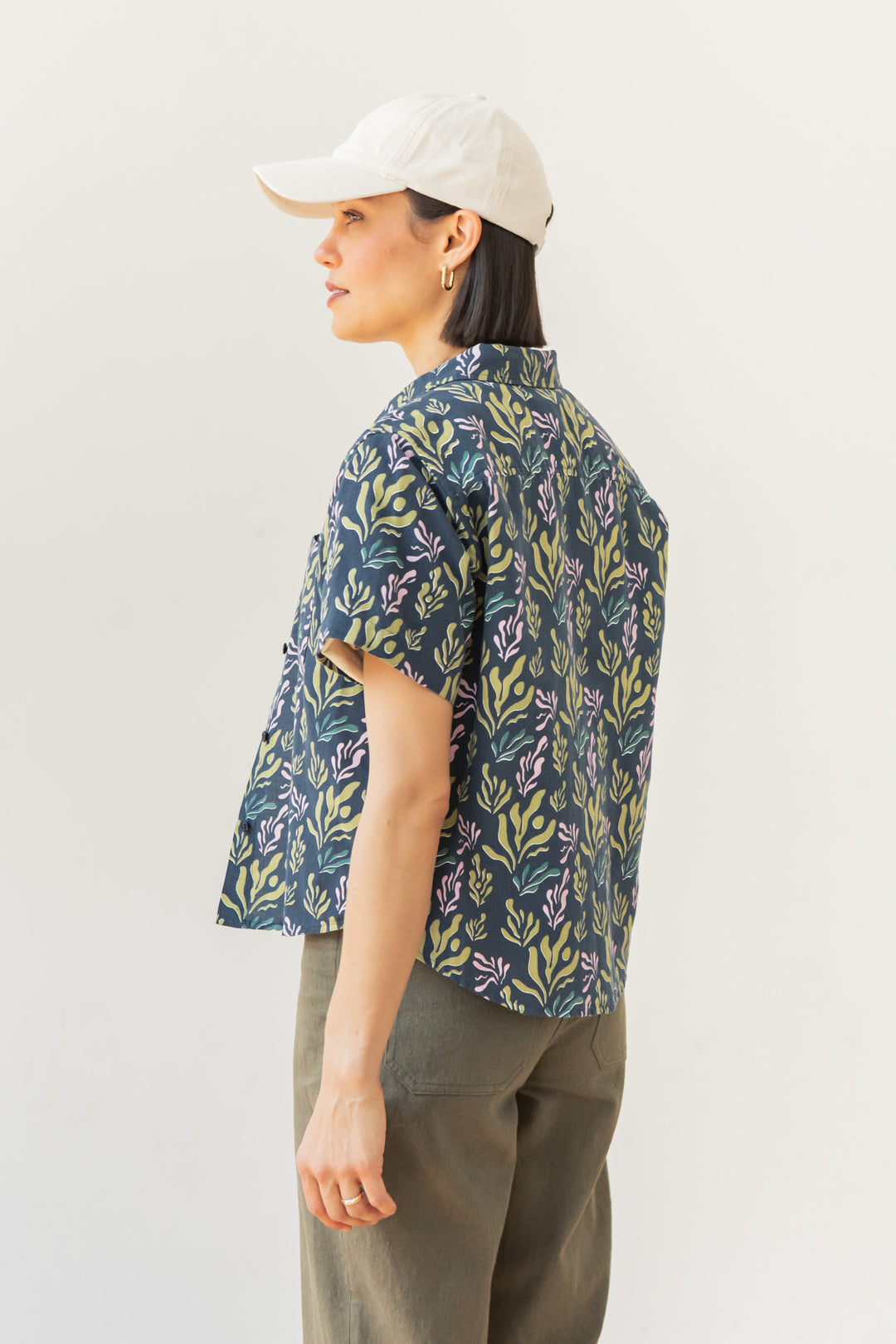 A person wearing the Bridge & Burn Greer Cropped Shirt in Spring Kelp, featuring a leafy design, paired with olive green pants and a white baseball cap, stands sideways against a plain white background.