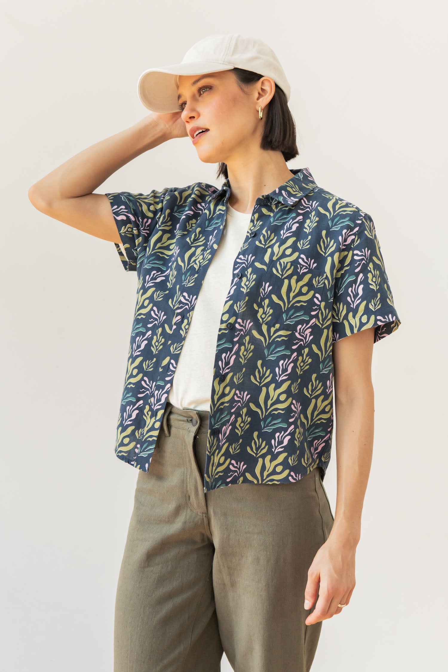 A woman wears the Bridge & Burn Greer Cropped Shirt in Spring Kelp, paired with a white t-shirt, olive pants, and a white baseball cap. She stands against a light background, touching her cap and looking to the side.