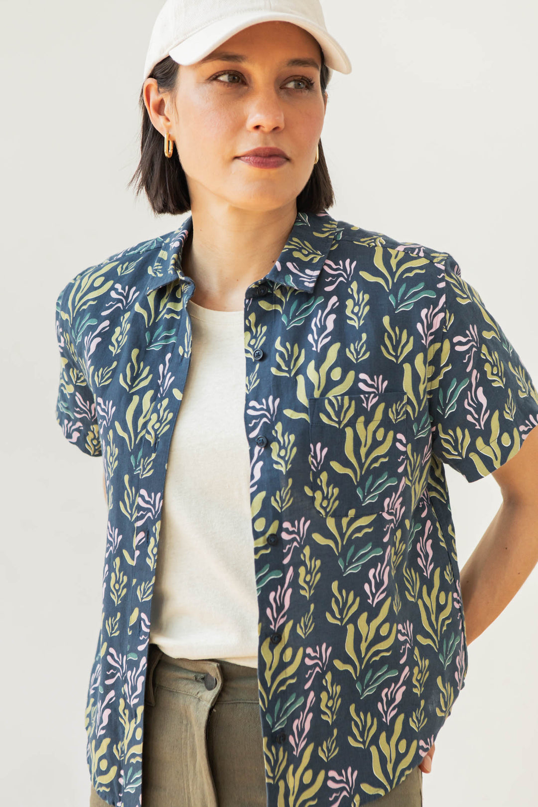 A woman wears the Bridge & Burn Greer Cropped Shirt in Spring Kelp over a white tee and white baseball cap, standing with one hand on her hip and looking to the side.