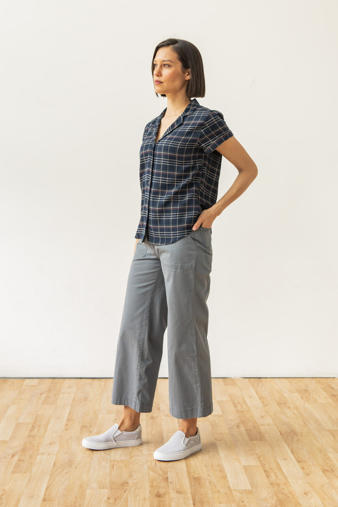 A woman with short dark hair stands indoors on a wooden floor, wearing the Bridge & Burn Innes Shirt in Dusk Herringbone Plaid, gray wide-leg pants, and white slip-on shoes. One hand is in her pocket as she looks forward.