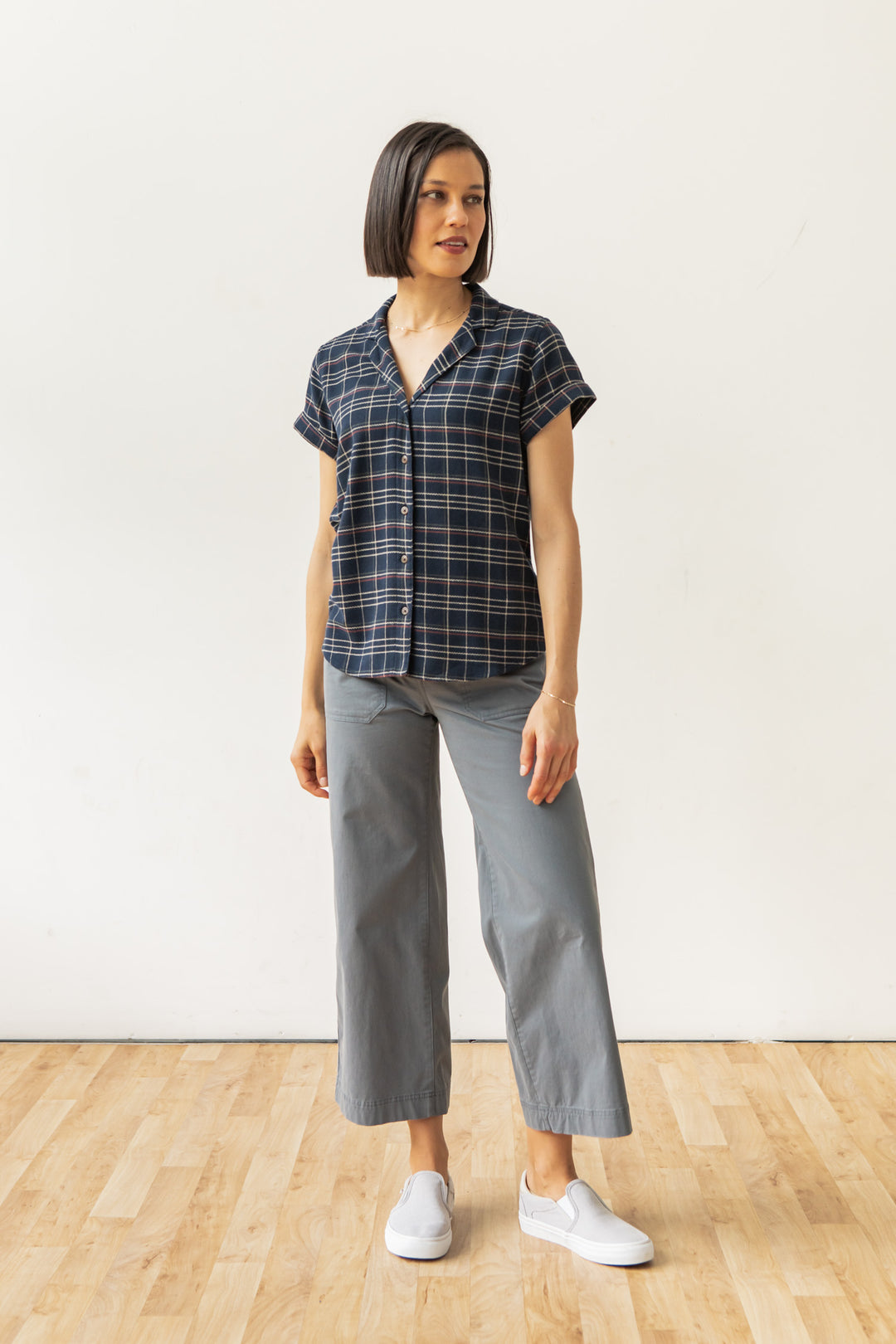 A woman stands on a wooden floor against a plain white wall, wearing the Bridge & Burn Innes Shirt in Dusk Herringbone Plaid, paired with gray wide-leg pants and white slip-on shoes. She looks slightly to her left.